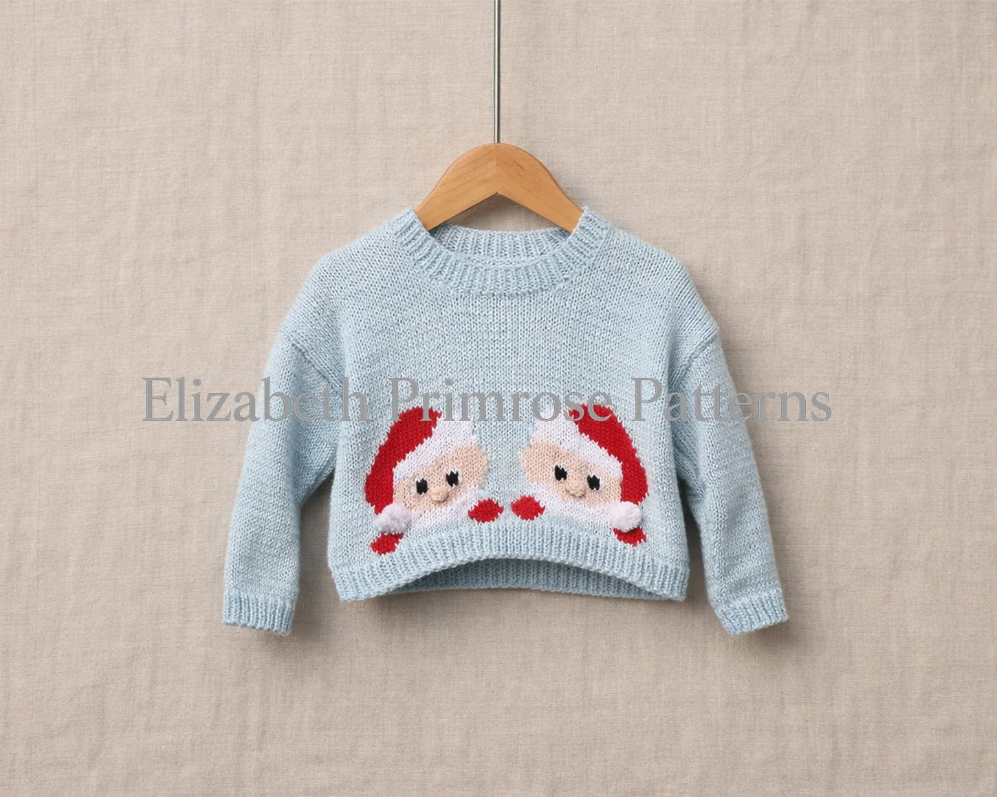 Light blue children’s jumper on a wooden hanger, featuring two peeking Santa motifs against a soft beige fabric background.