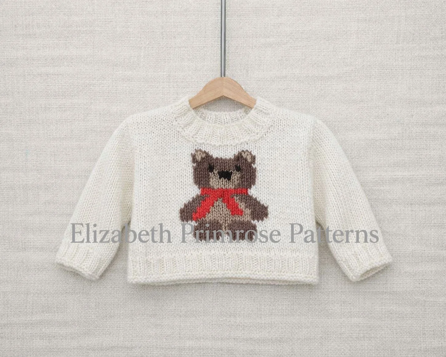 Cream knitted children’s jumper with brown teddy bear and red scarf motif, hanging on a wooden hanger against a beige fabric background.