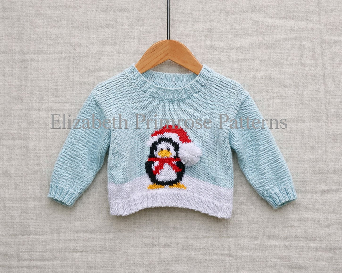 Light blue children’s jumper with penguin in Santa hat displayed on a hanger against a soft beige fabric background.