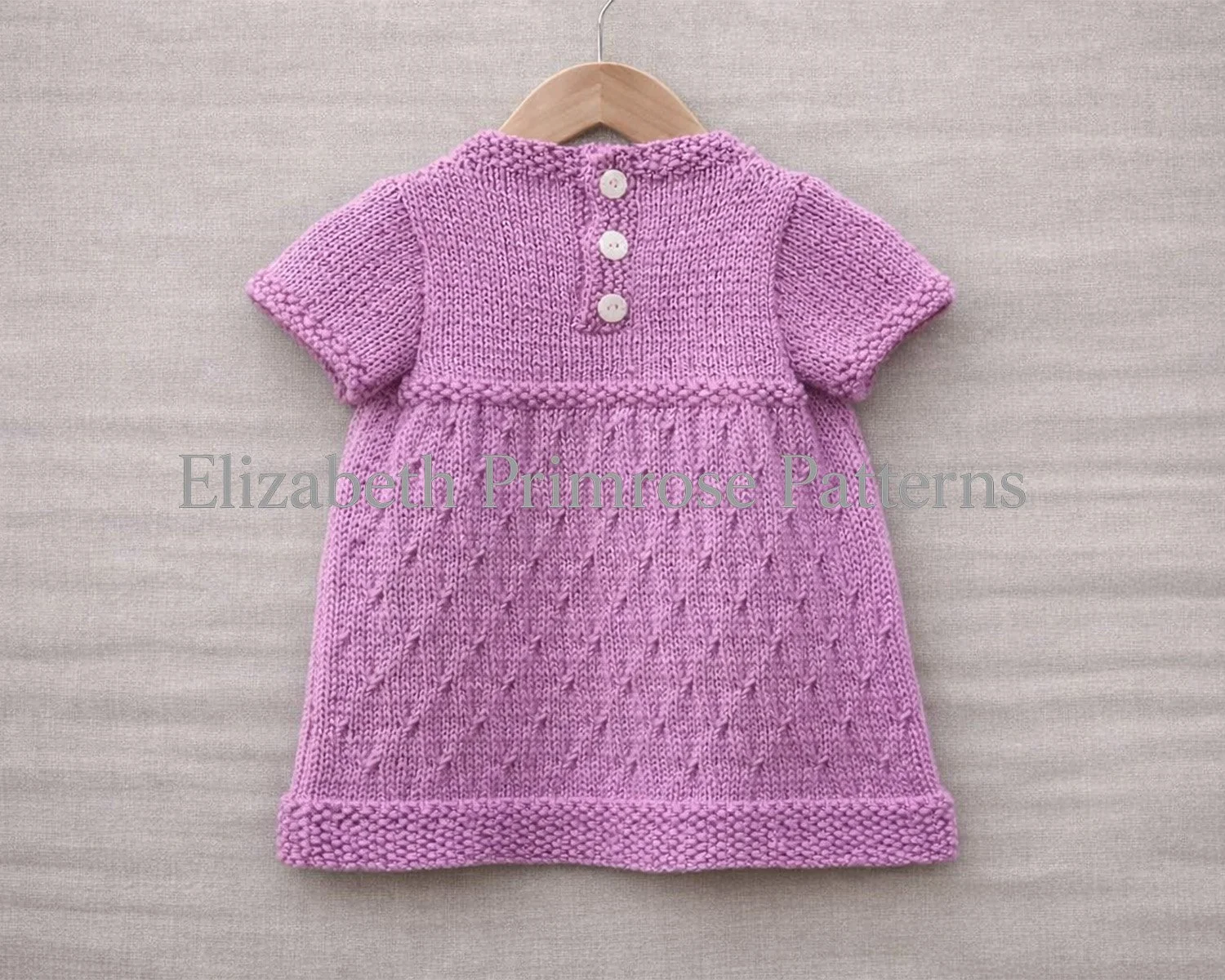 Back view of knitted pink dress on a wooden hanger against a soft linen background.  The dress has a 3 button band and a textured skirt.