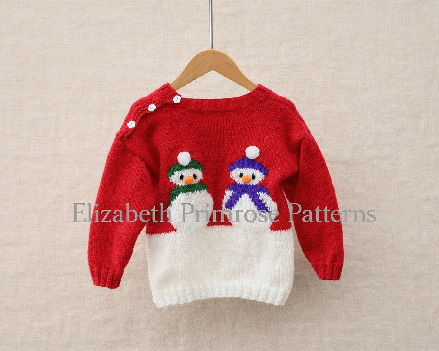 Red and white hand knitted children’s jumper with two snowmen wearing green and blue hats, displayed on a wooden hanger against a beige linen background.