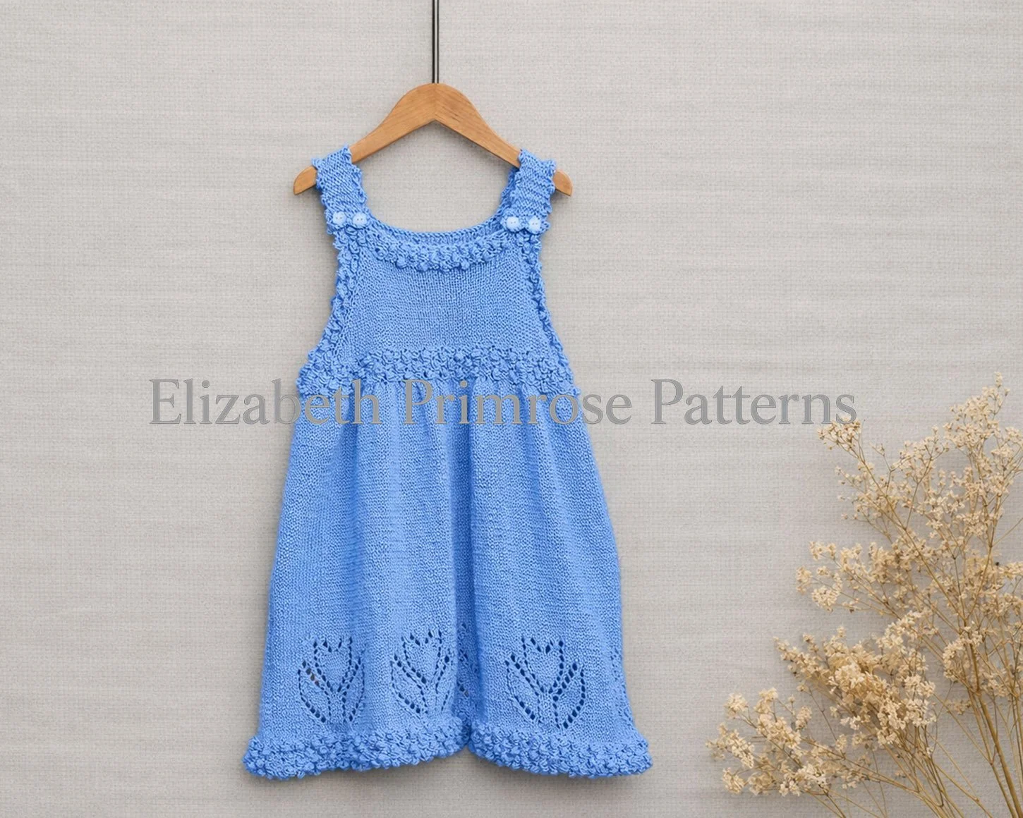 Light blue hand knitted dress with tulip and bobble hem pattern hanging on wooden hanger against beige background beside dried flowers.