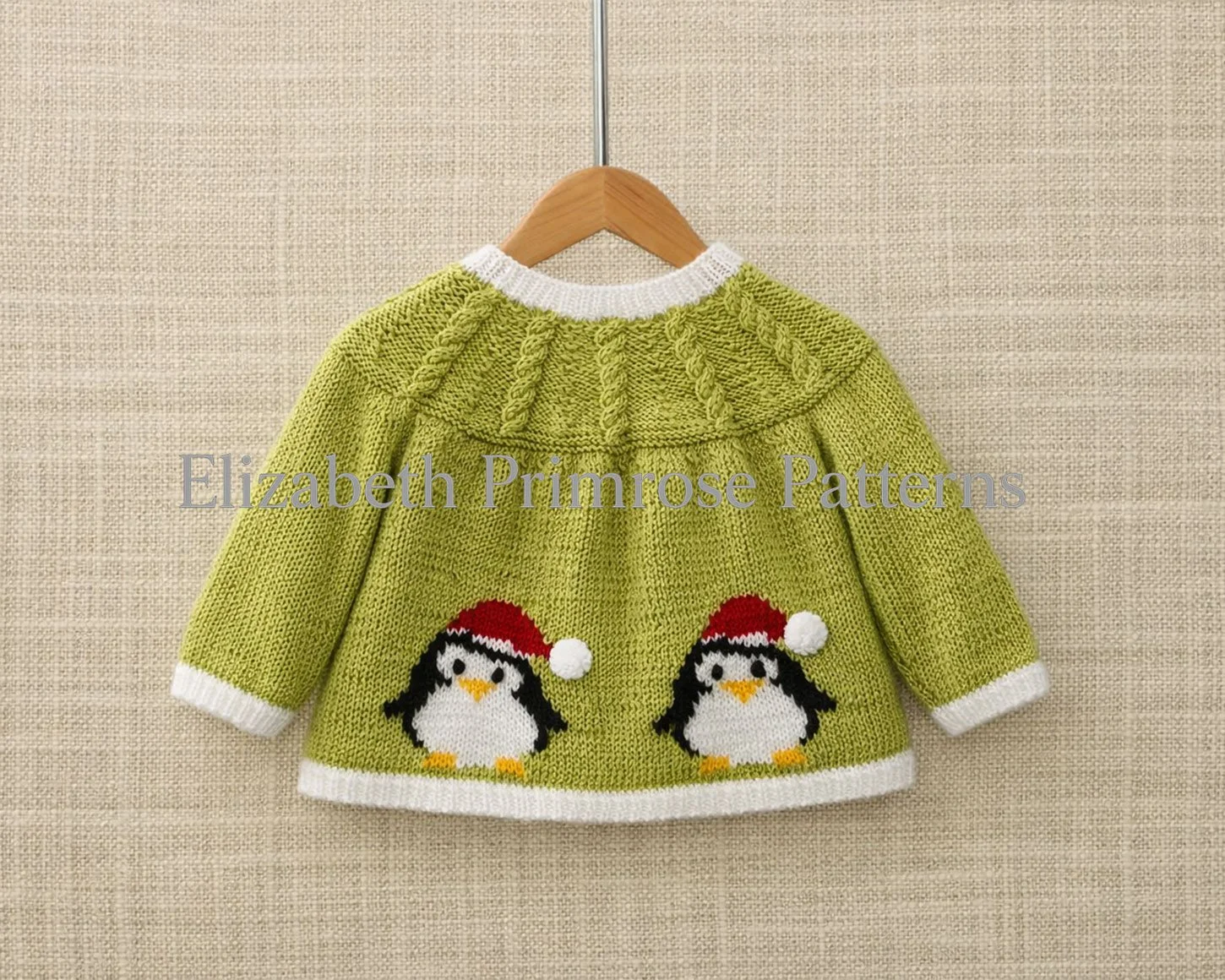 Back of green knitted baby cardigan with penguin motis, white trim and cabled yoke, hanging on a wooden hanger against a beige linen background.