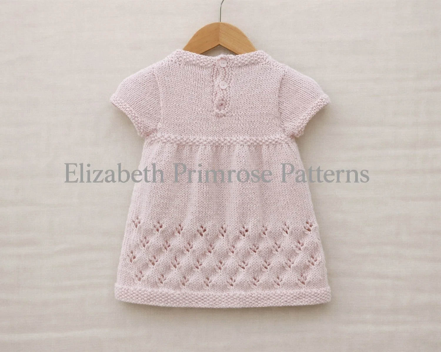 Back view of a pale pink hand knitted baby dress with short sleeves, hanging on a wooden hanger over an uncreased light beige linen background.