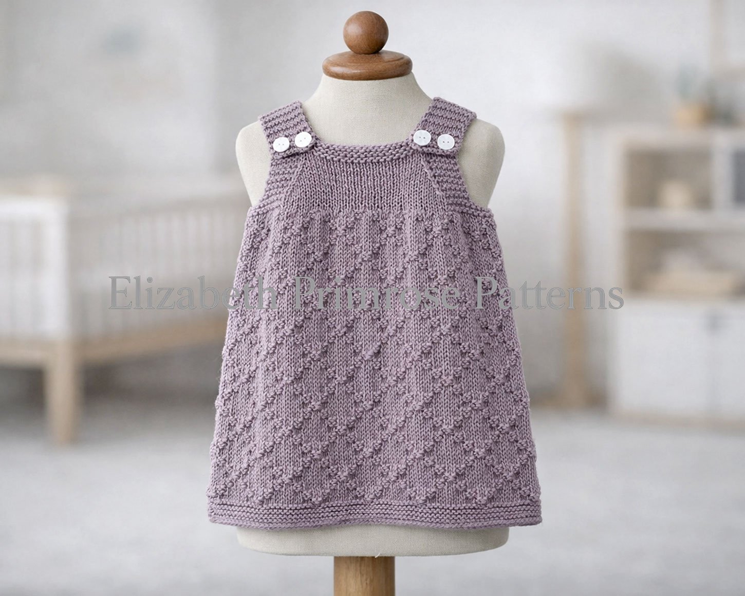 Textured lavender baby pinafore dress with white buttons shown on a mannequin in a gentle nursery setting. Diamond lattice design. Elizabeth Primrose Patterns knitting pattern.