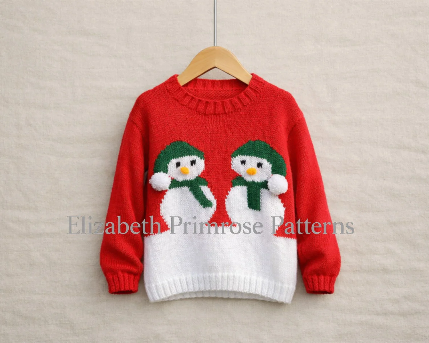 Red and white children’s jumper on a wooden hanger, featuring two snowmen with green hats and scarves on a soft beige linen background.