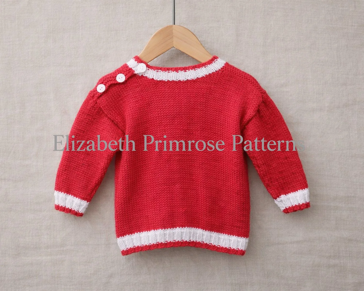 Back of red knitted baby sweater with white ribbed collar, cuffs, and hem, hanging on a wooden hanger with straight metal pole over beige linen.