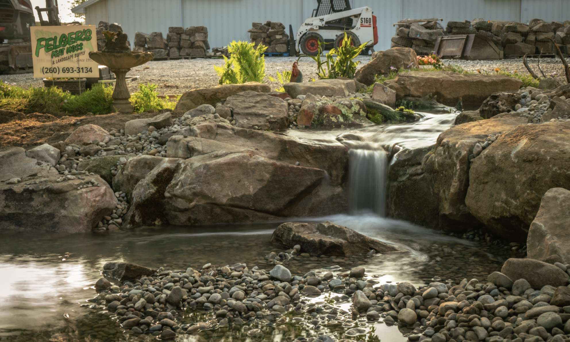 Decorative garden waterfall with rocks, plants, and a sign for Felger's Peat Moss & Landscape Supply in the background.