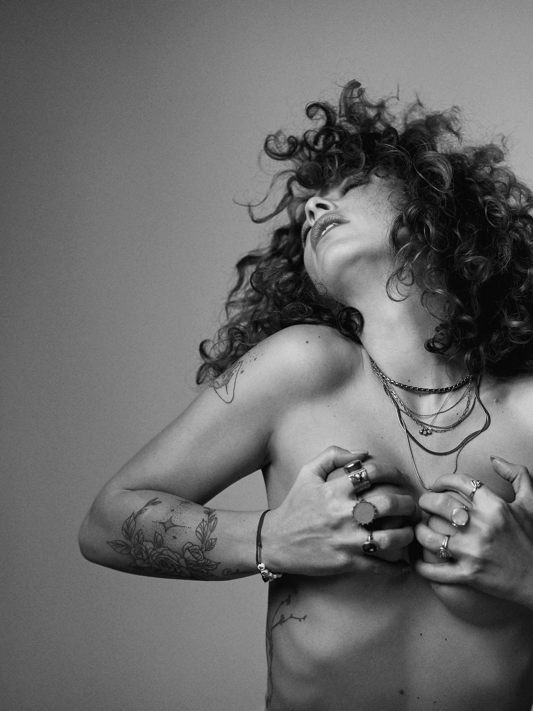 Une femme avec des cheveux bouclés, portant plusieurs bijoux, se couvre partiellement la poitrine avec ses mains, dans une photo en noir et blanc.