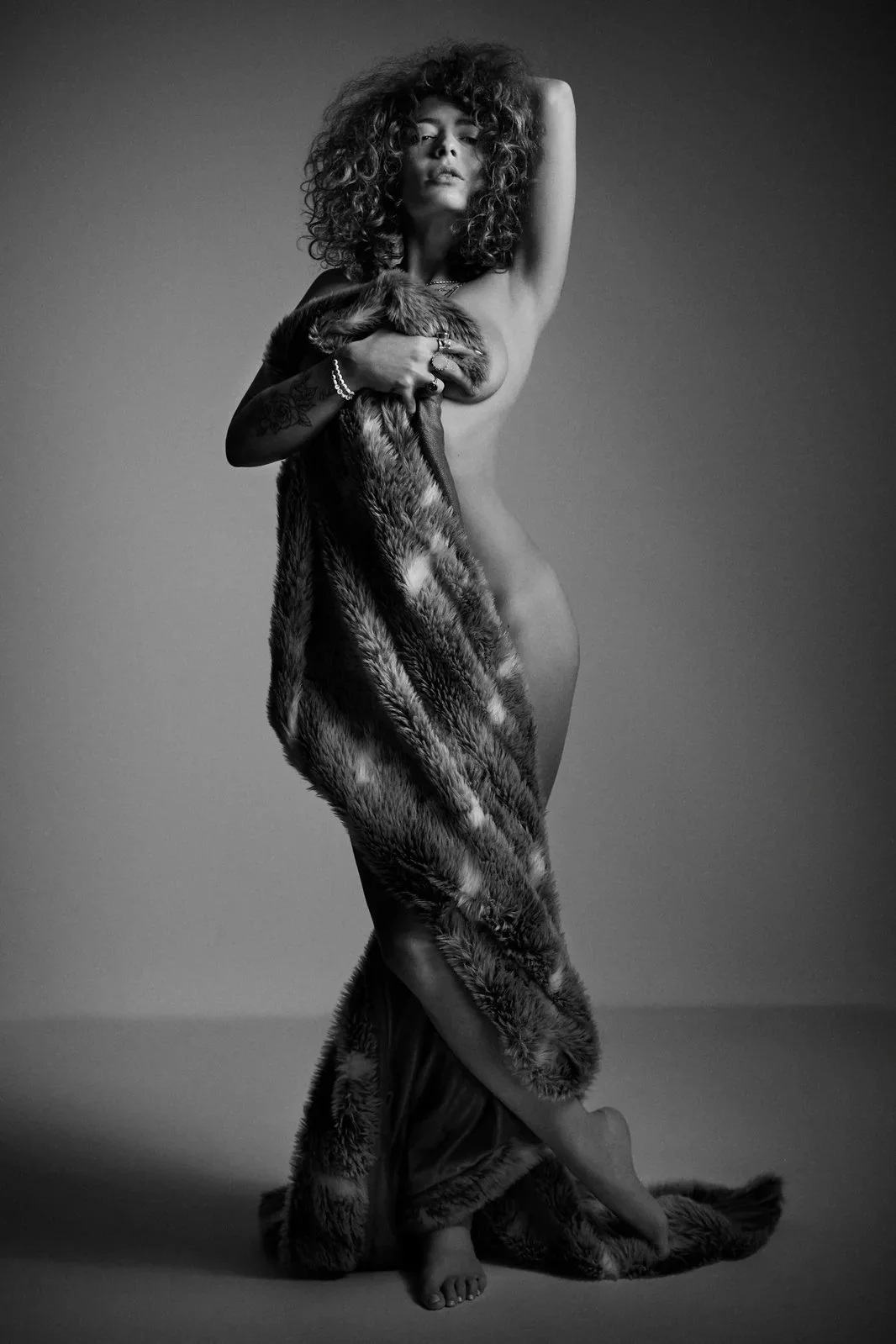 Une femme nue, avec des cheveux bouclés, se couvre partiellement avec une robe en fourrure, tenant un morceau également en fourrure, sur un fond gris foncé.