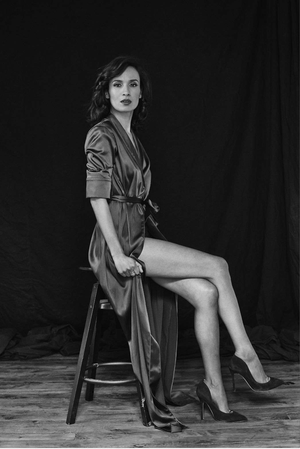 Une femme en robe de satin, assise sur une chaise, portant des talons hauts, avec un arrière-plan noir
