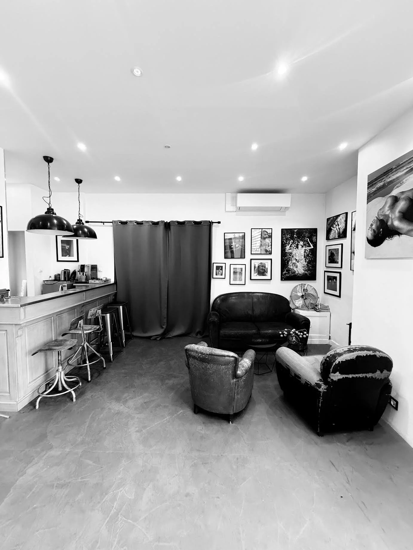 Salon intérieur avec un mur de cadres, un canapé en cuir noir, des fauteuils anciens, une table basse, et une petite table à côté, éclairage par plafonniers et luminaires suspendus