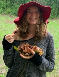 Young woman outdoors wearing a red hat and gray sweater, holding a plate with food and smiling.