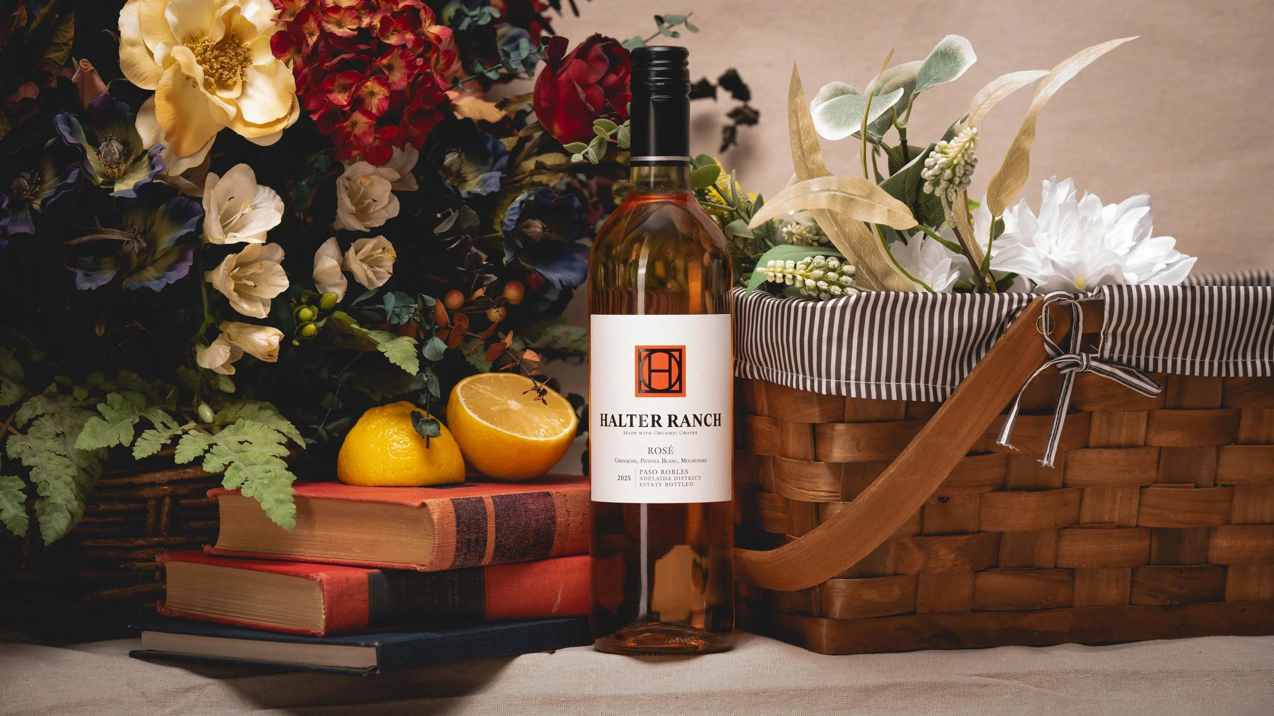 A table arrangement featuring a bottle of Halter Ranch rosé wine, a basket of white and dark flowers with green foliage, a halved lemon, and two hardcover books, all set on a tablecloth.