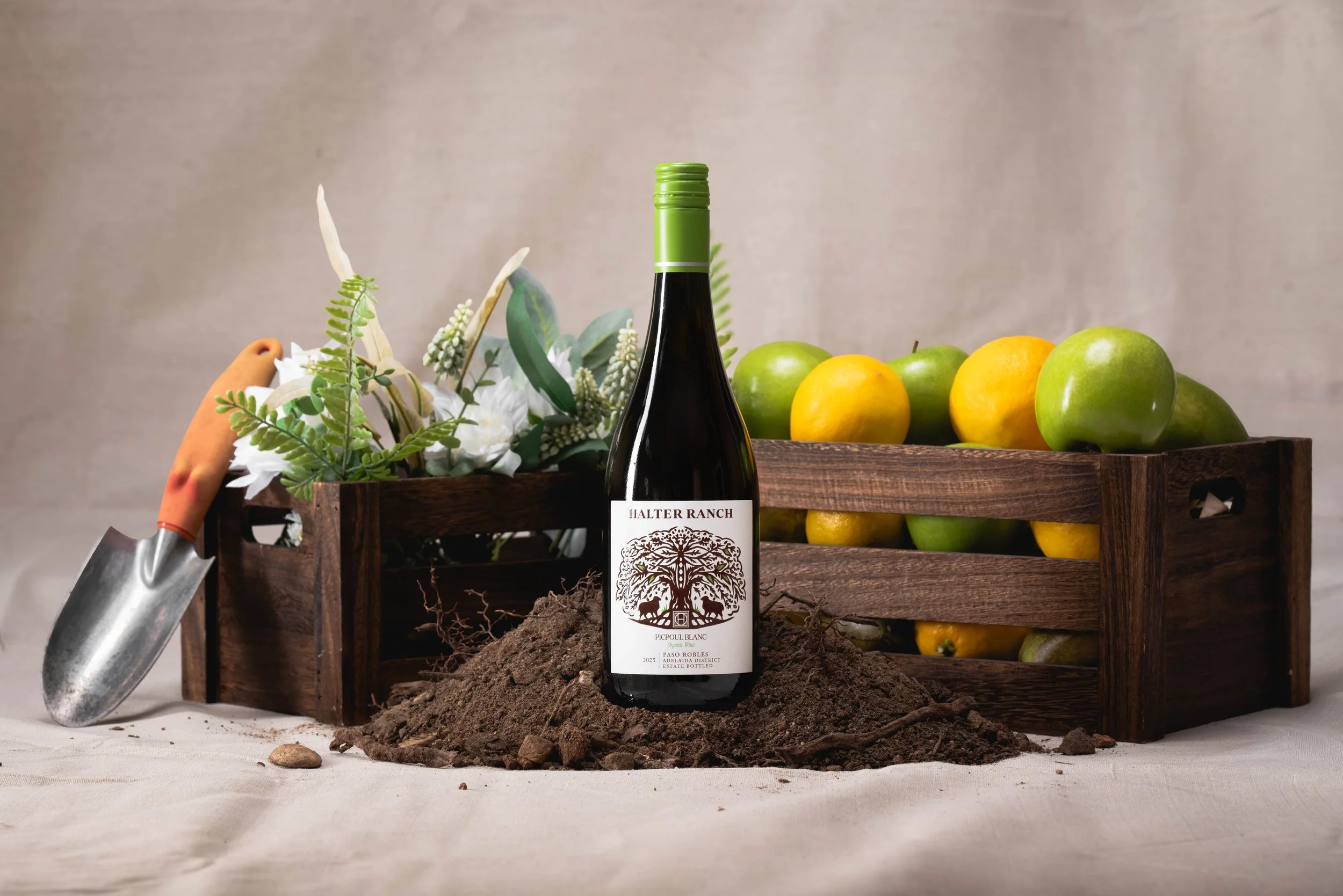 A bottle of Halter Ranch wine in front of a pile of dirt, with green and yellow lemons in a wooden crate, a garden trowel, and decorative greenery and flowers in the background.