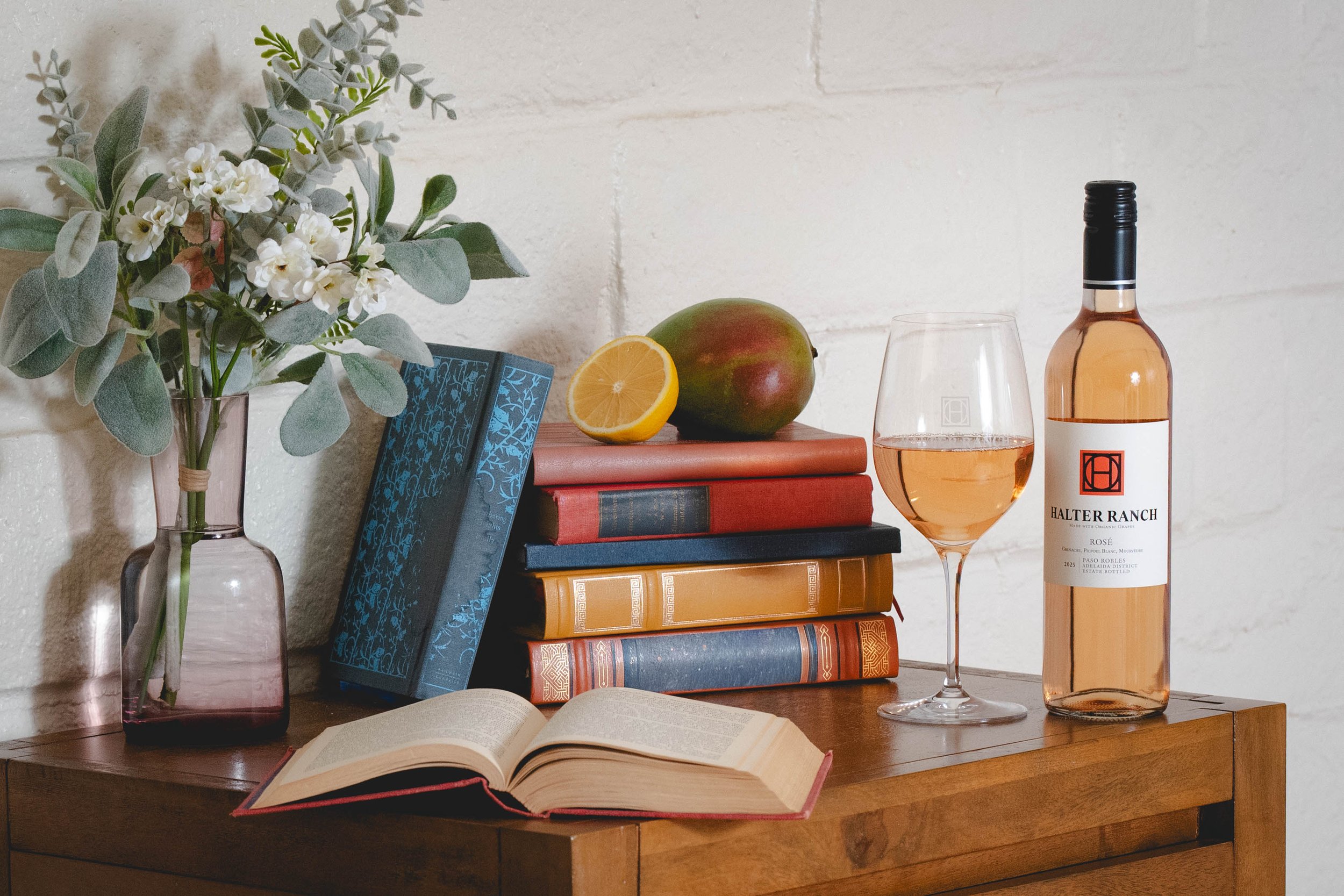 A wooden table displaying a vase with flowers, stacked books with a mango and lemon on top, an open book, a glass of rosé wine, and a bottle of Halter Ranch rosé wine in front of a white brick wall.