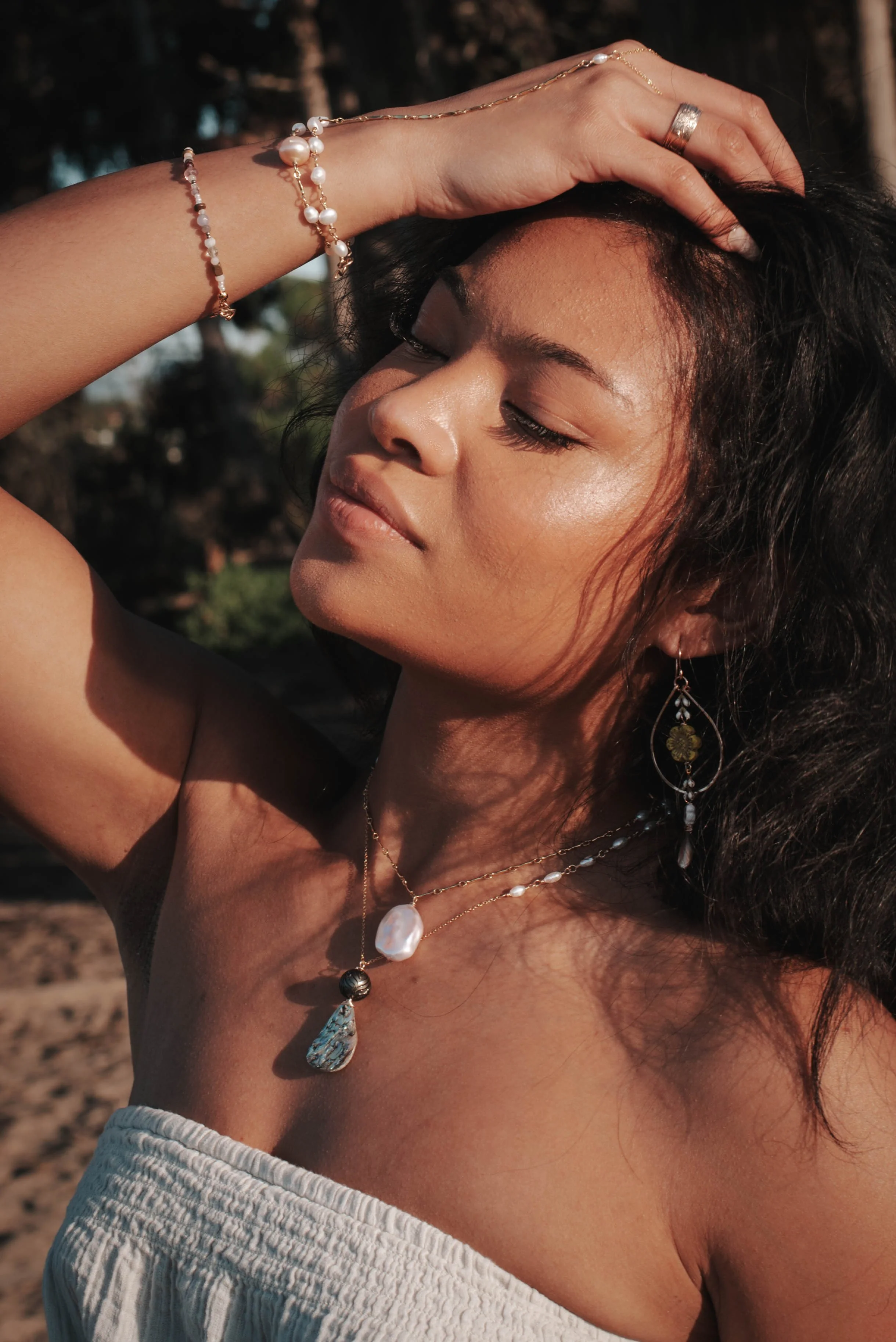 A woman with dark, wavy hair wearing jewelry, including layered necklaces and earrings, with her hand resting on her head and eyes closed.