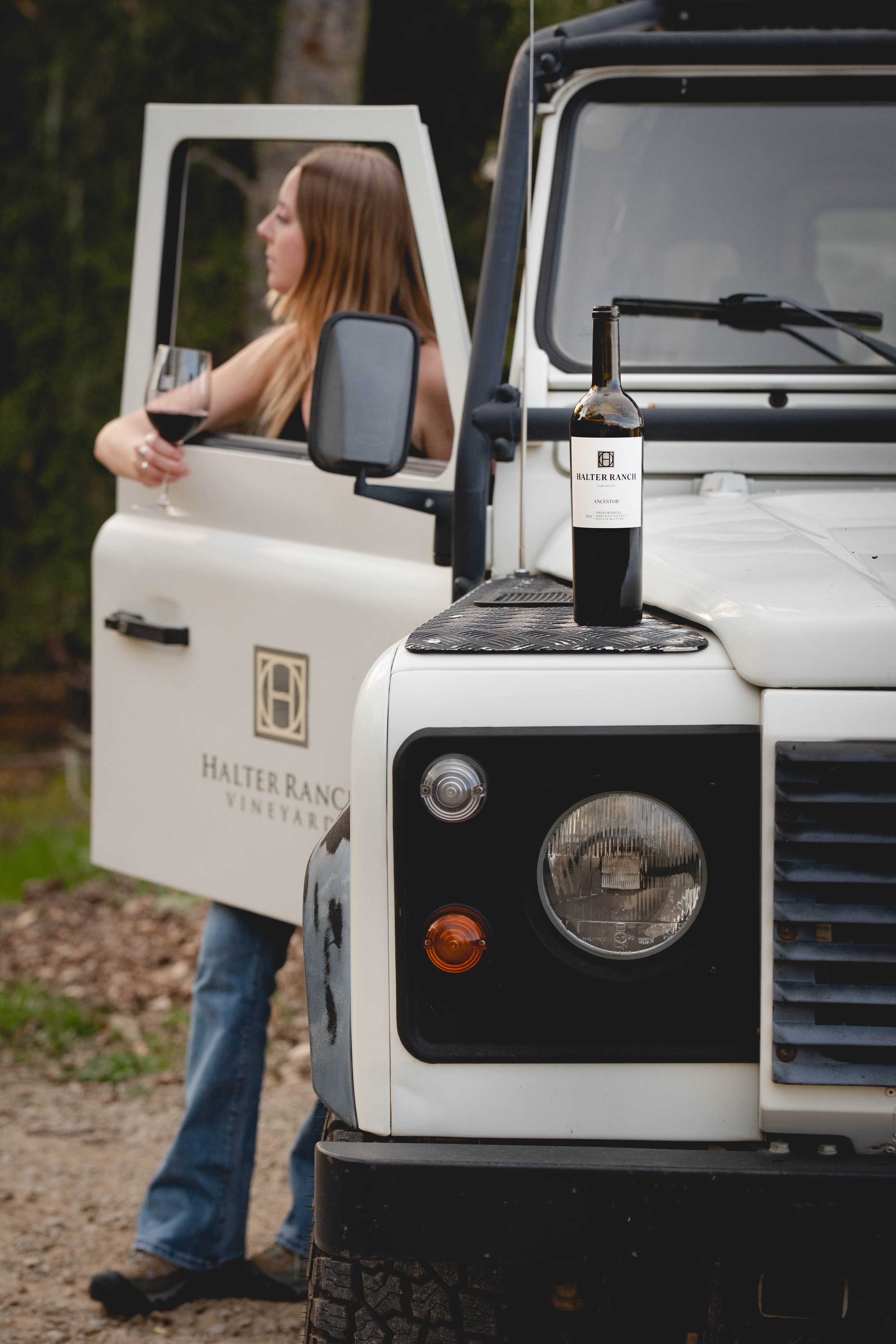 A woman with long blonde hair holding a glass of red wine standing outside next to a white utility vehicle with a bottle of wine on the hood, vineyard logo and landscape trees in the background.