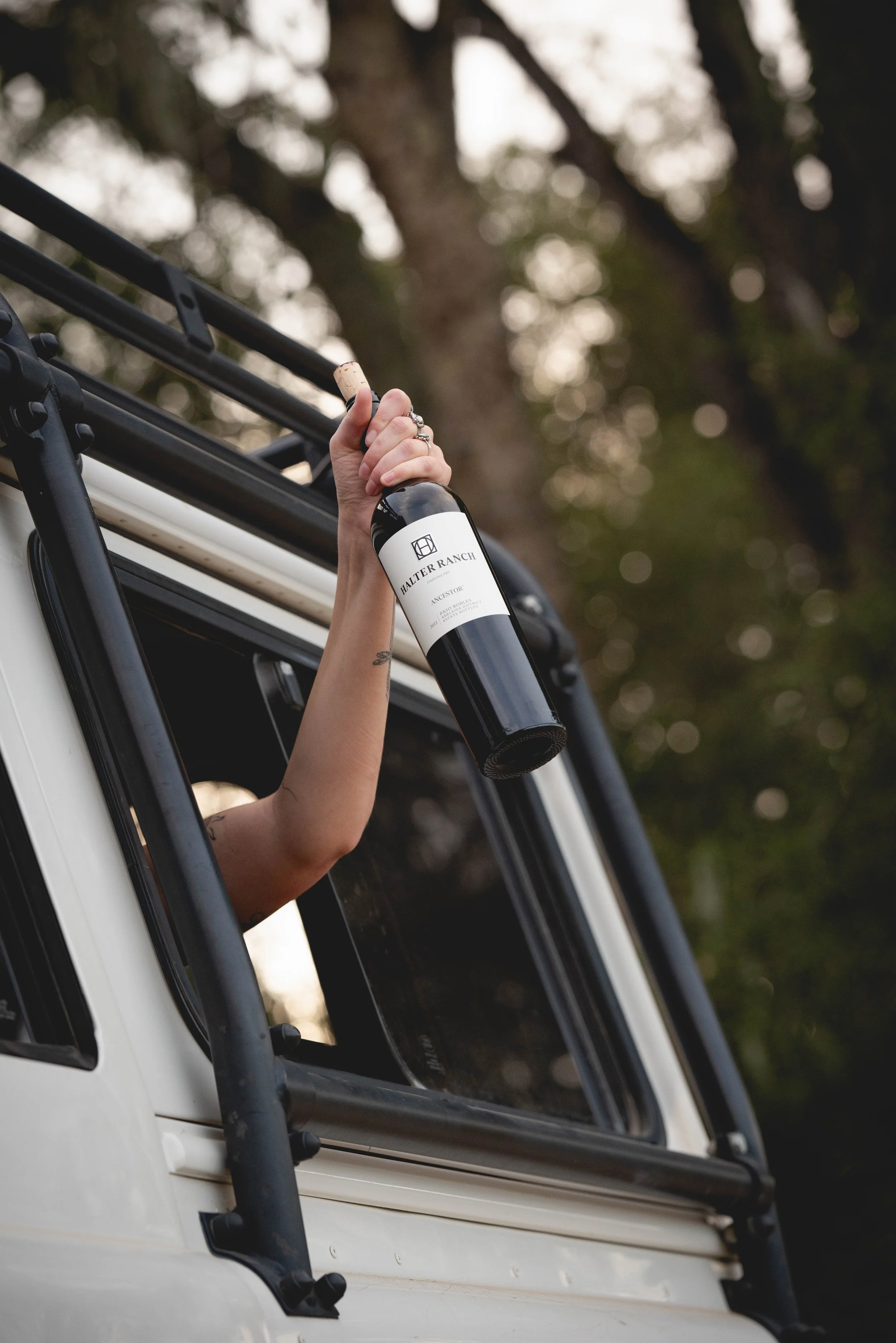 Person holding a bottle of red wine labeled 'Walter Ranch Accistor' out of a vehicle window with a wooded background.