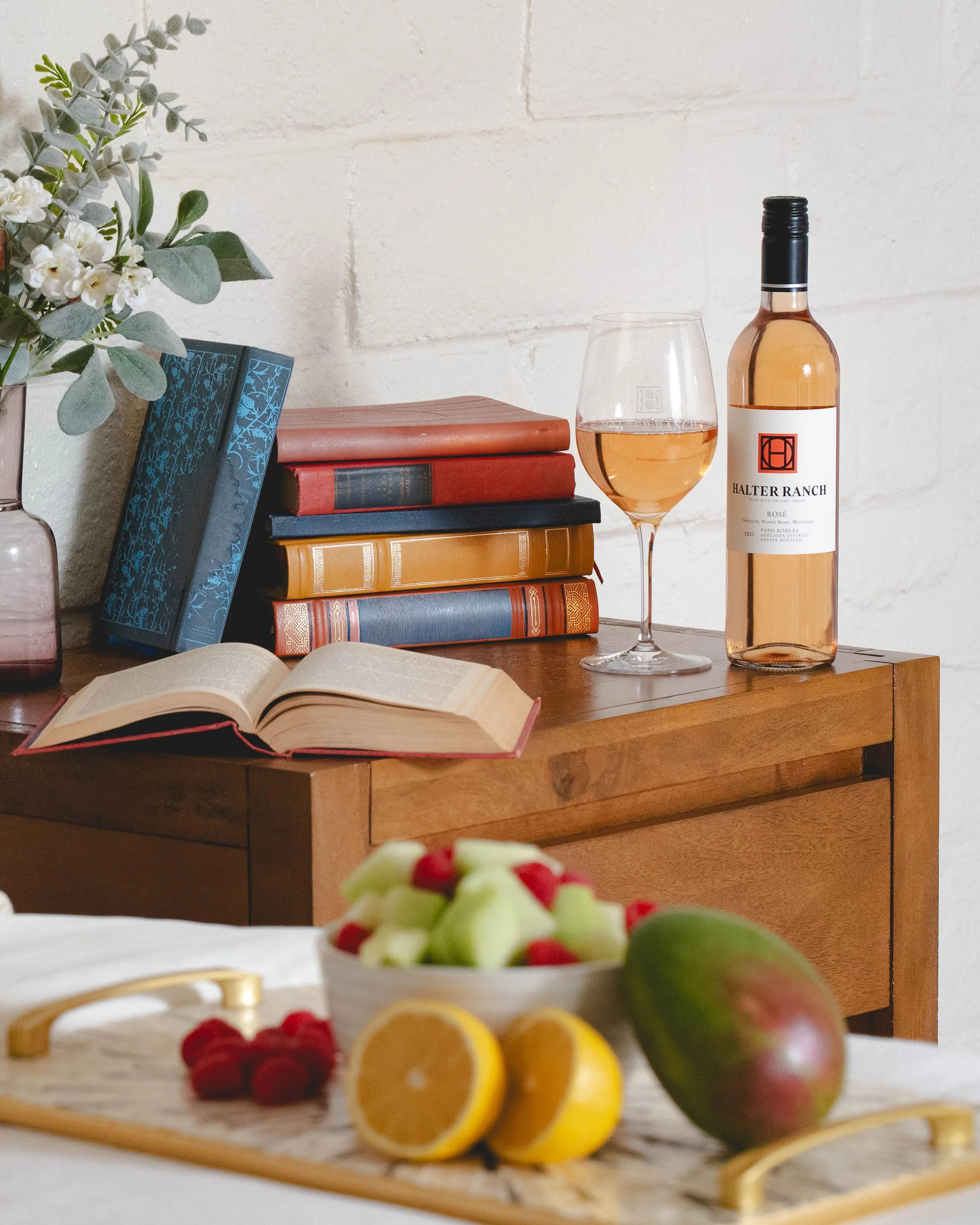 A wooden nightstand with a pile of books, an open book, a glass of rosé wine, a bottle of rosé wine, and a bowl of fresh fruit including melon, raspberries, lemon, and a mango.