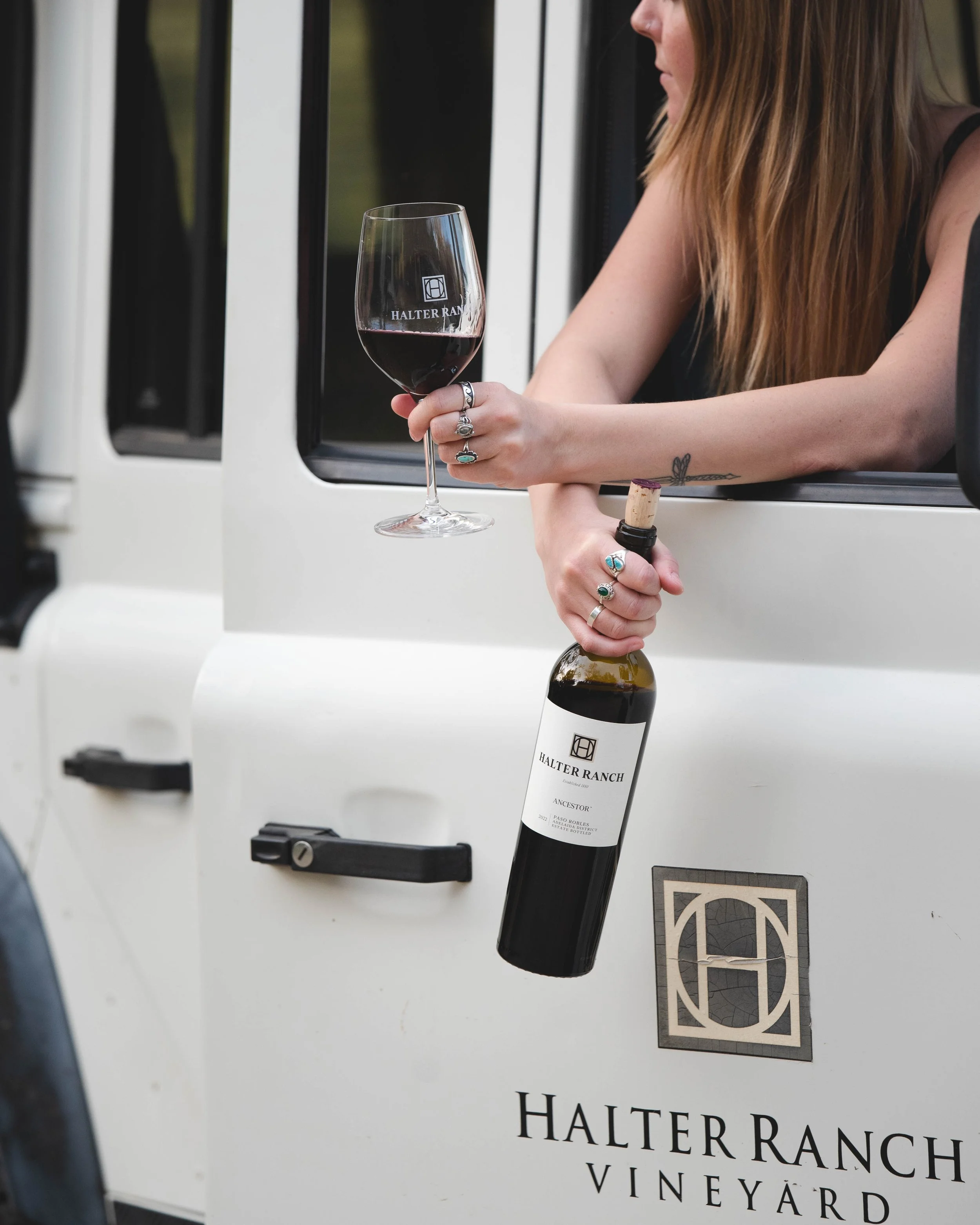 Person with long hair holding a glass of red wine and a bottle of wine while leaning out of a white vehicle with 'HALTER RANCH VINEYARD' logo and text.