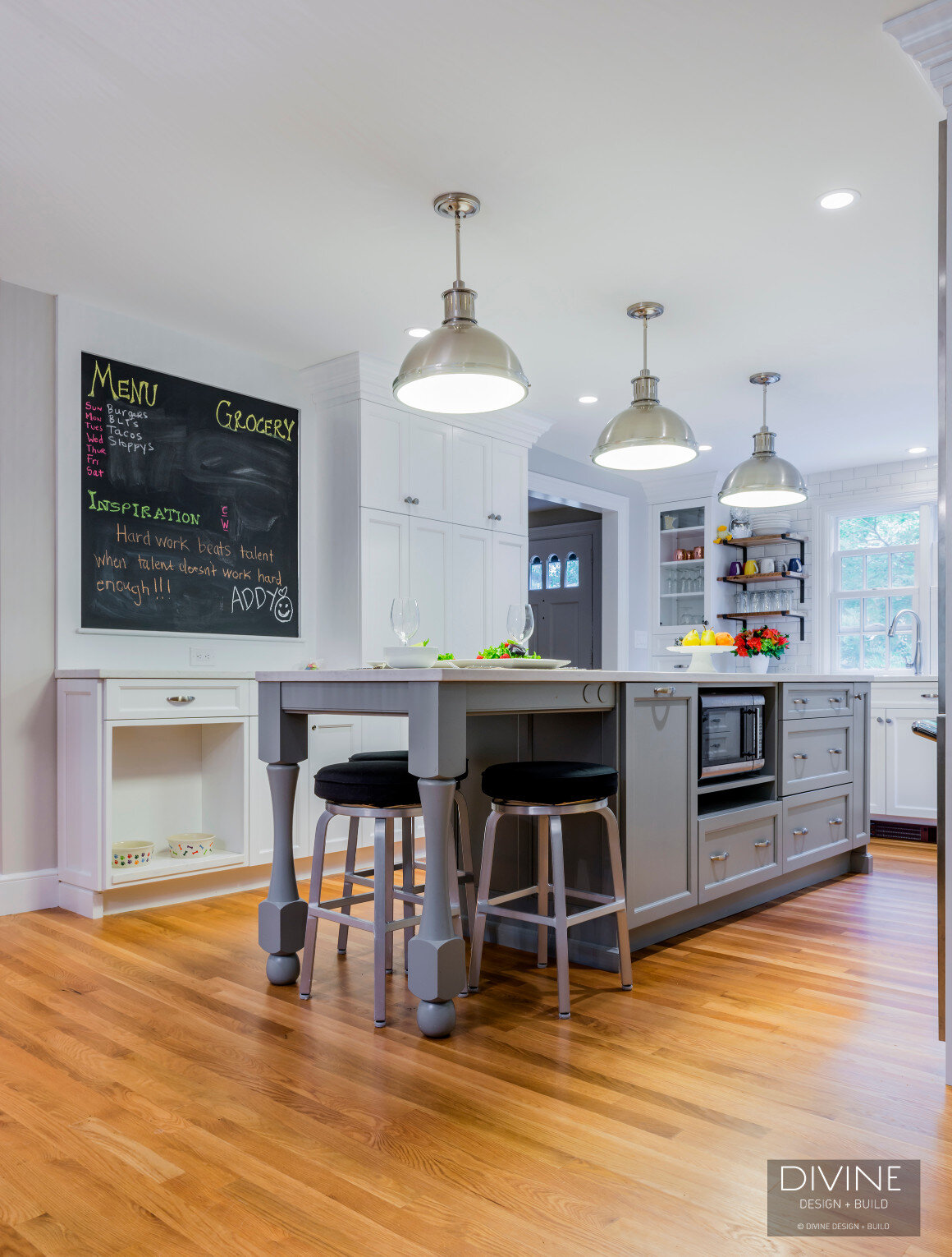  Grey and white transitional style kitchen with shaker cabinets and integrated dog feeding station. Chalkboard. Subway tile backsplash and industrial style lights and shelving