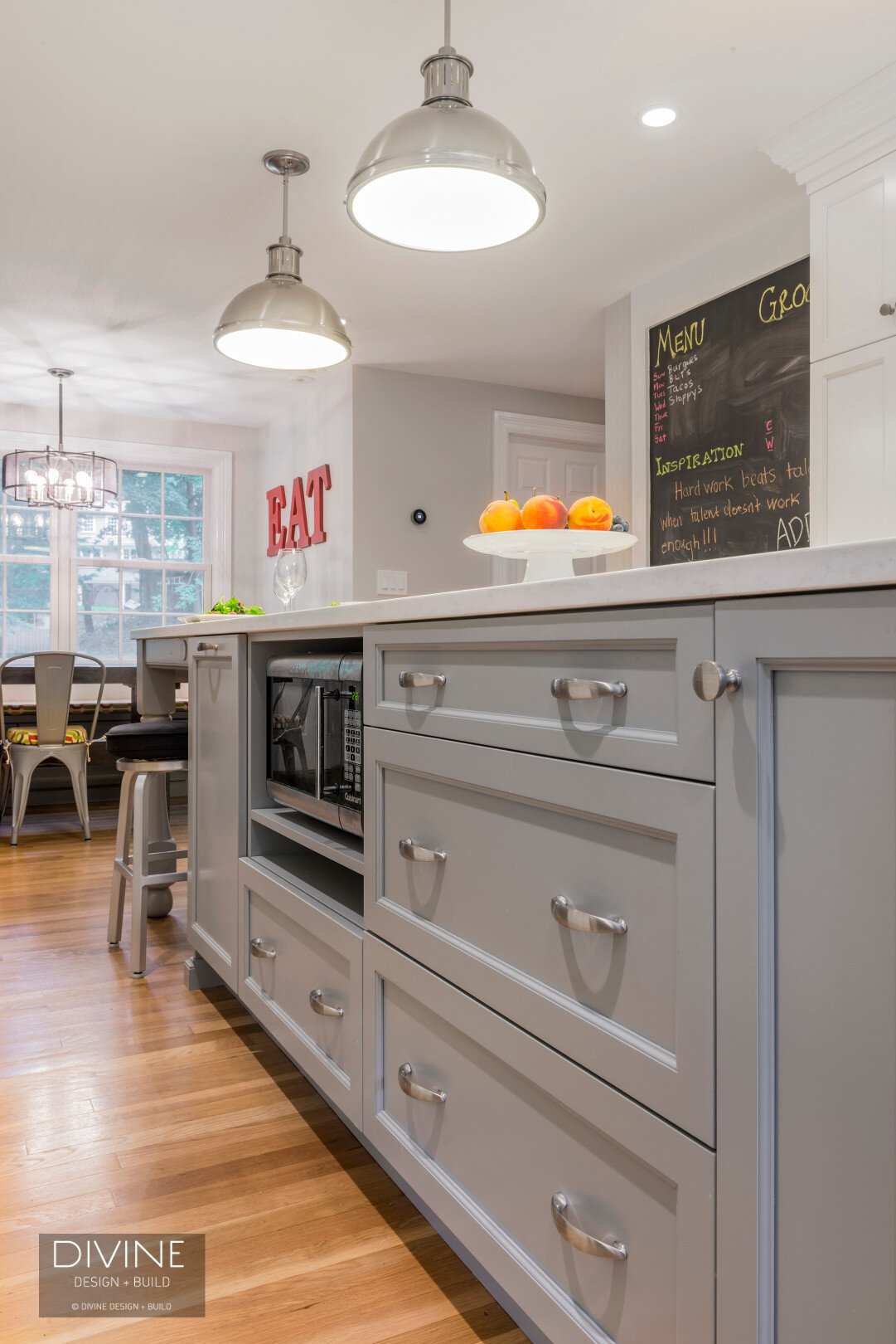 Grey and white transitional style kitchen with shaker cabinets and integrated dog feeding station. Chalkboard. Subway tile backsplash and industrial style lights and shelving
