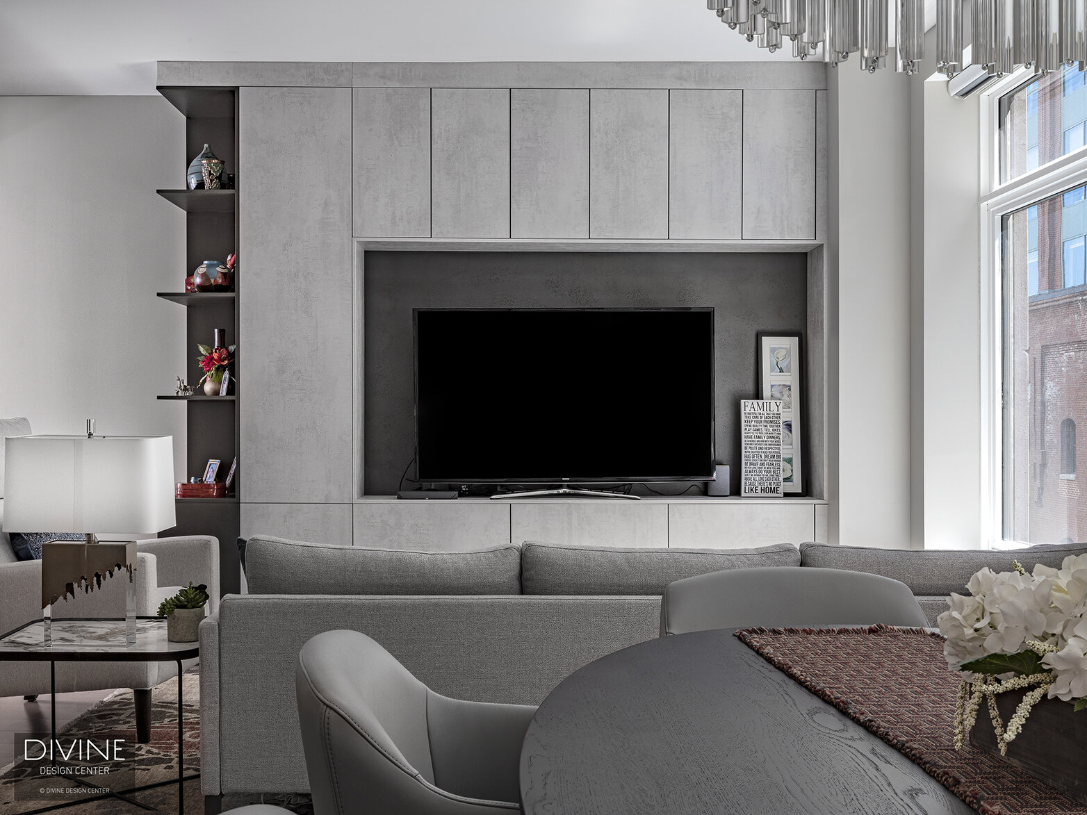   Faux concrete built-ins surround a flat-screen tv. Niche shelving is installed to the left of the tv surround and is adorned with several objects and vessels on display. Rolf Benz dining chairs and a round, wood dining table sit in the corner of th