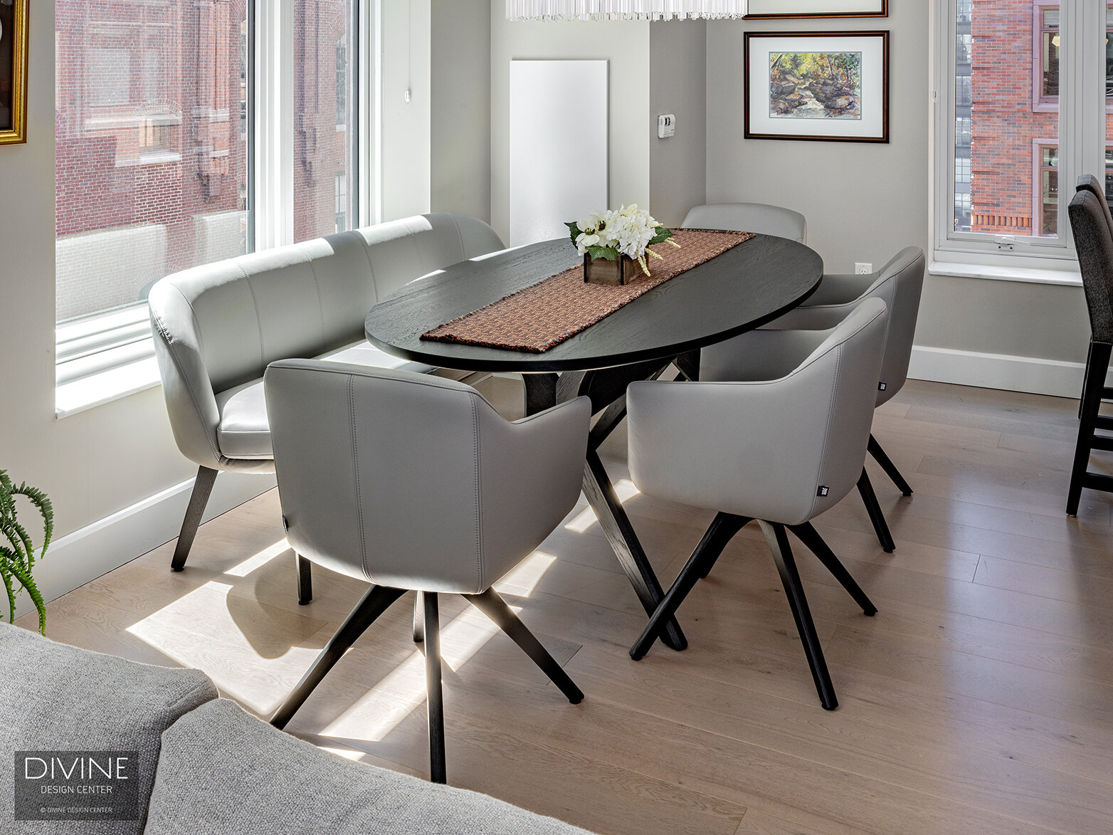  Rolf Benz dining chairs and a bench sit around a round, wood dining table in the condominium’s dining area. Large windows cast a bright, sunny light over the area.  