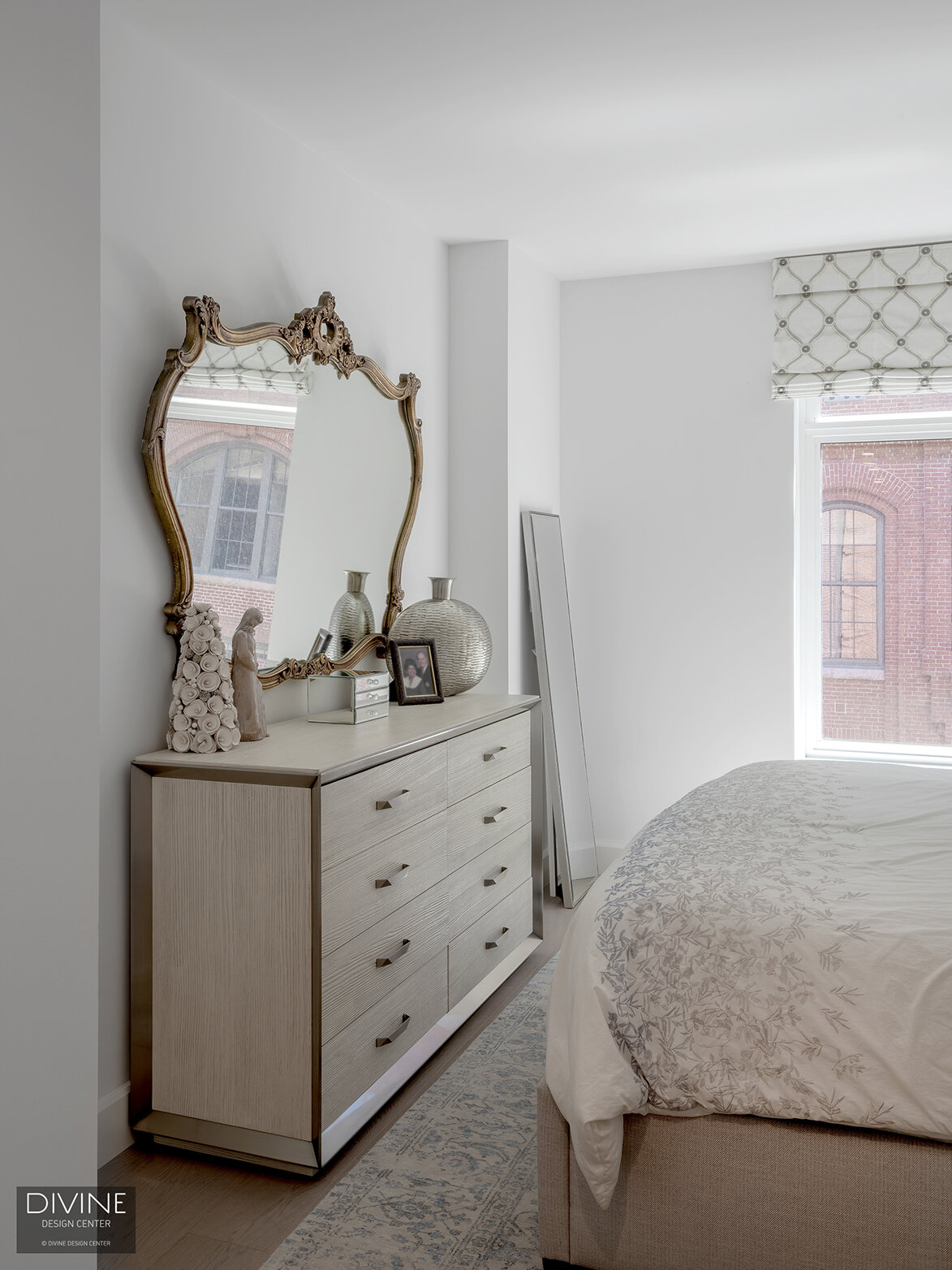  An ornate mirror and transitional style drawer system sit in front of an upholstered bed frame with delicate sheets adorning the mattress. 
