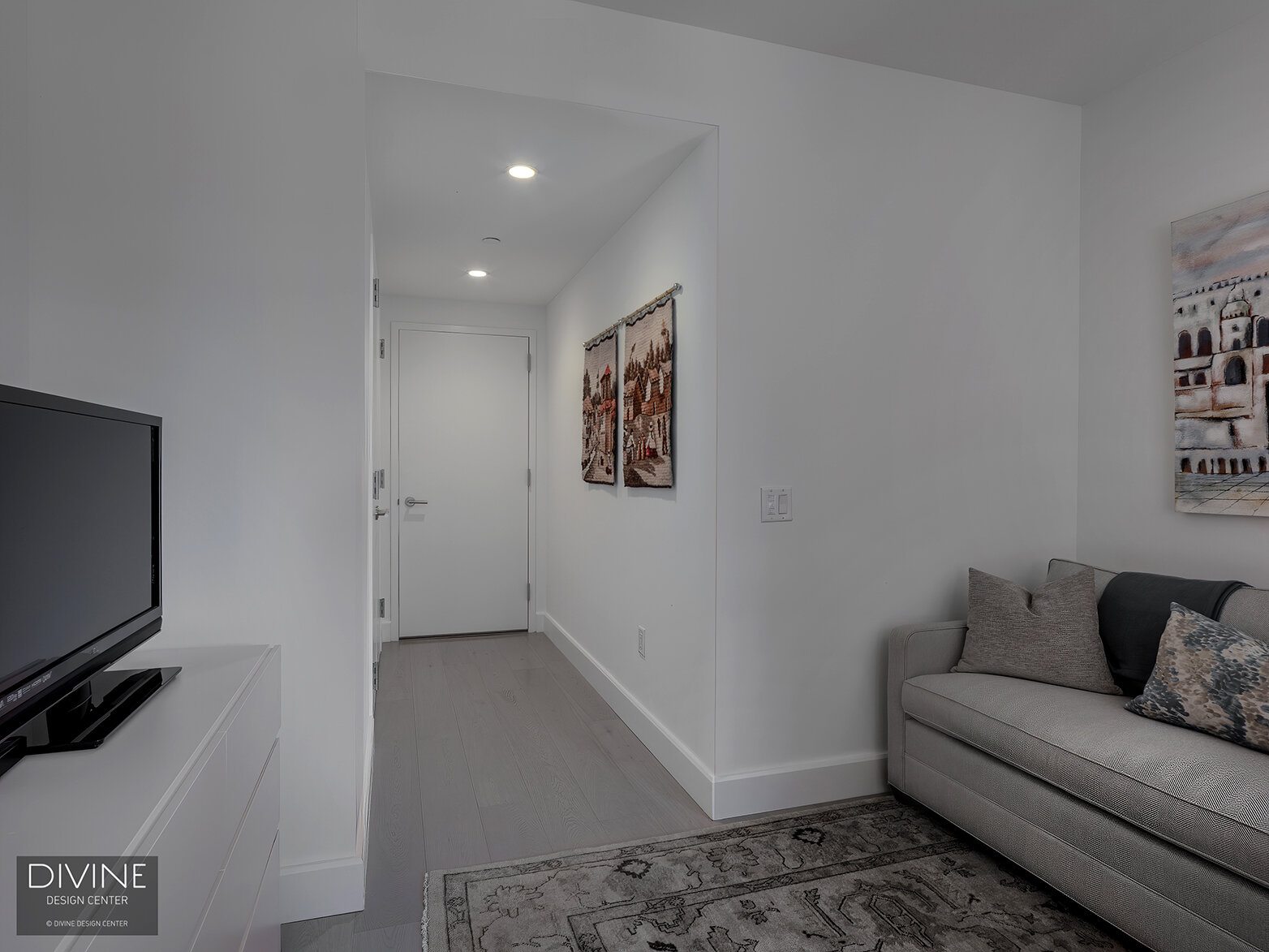  A modern home office with a modern desk and Eames office chair is flanked by a modern, grey love seat sofa and white, handleless cabinets with a small tv sitting on top. A rug fills the room and one large window looks out onto the surrounding buildi