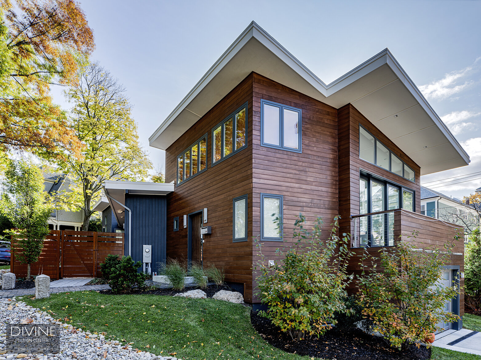  Contemporary home exterior with modern dark wood siding, black window frames, and a flat, architectural corrugated roof is surrounded by a large green lawn and healthy shrubbery.  