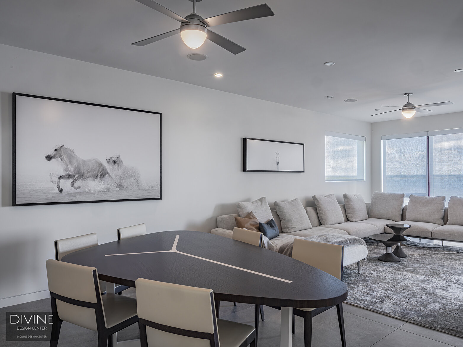  Open concept living/dining space with ocean views. An architectural dining room table accompanies a large grey sectional sofa with modern art hanging on the white painted walls. A large grey, textural rug sits below the sofa.   