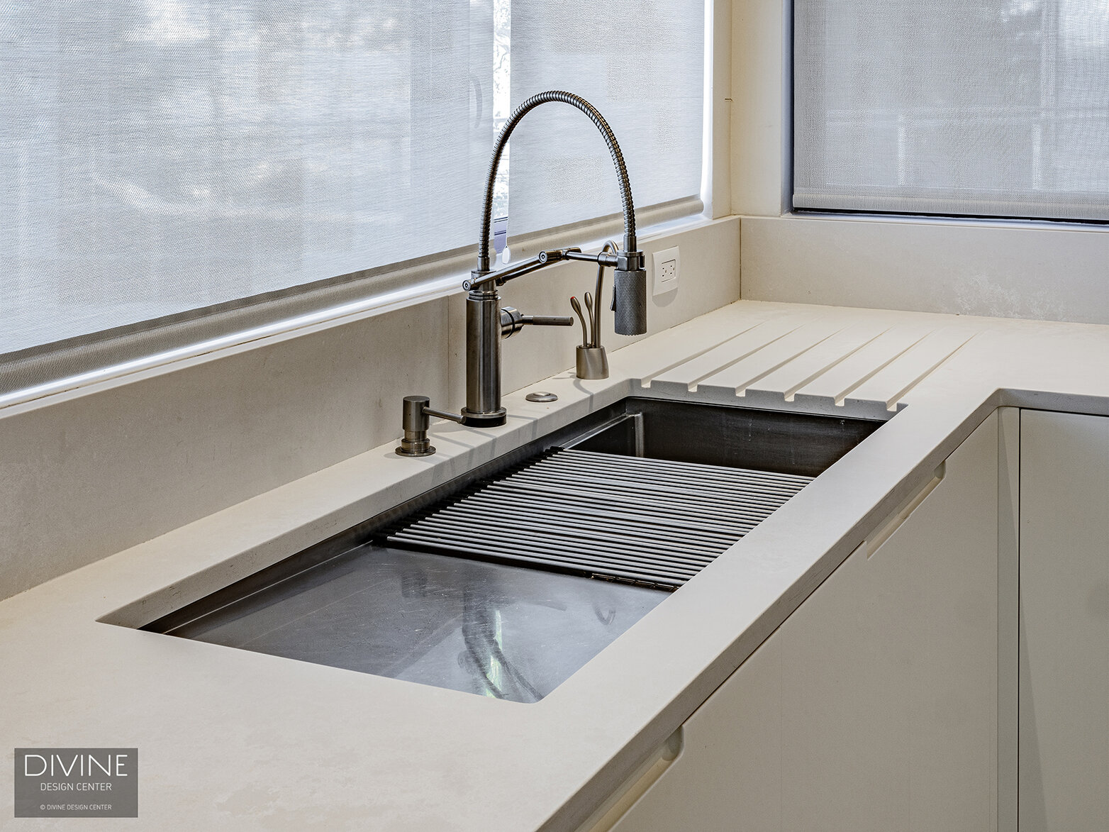  Light stone kitchen counter with a recessed, stainless steel sink. A custom-designed kitchen drying rack is debossed into the natural stone tops and complemented by a large oil-slicked kitchen faucet. Large windows looking out to the ocean are above