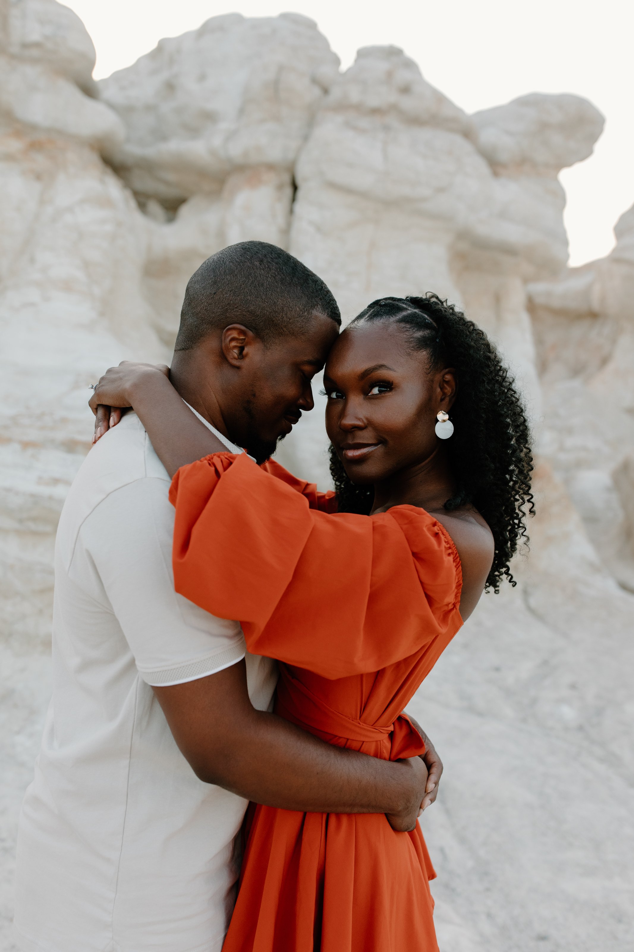 A couple standing close together in front of white rock formations, embracing with eyes closed and smiling, the woman wearing an orange dress and the man in a white shirt.