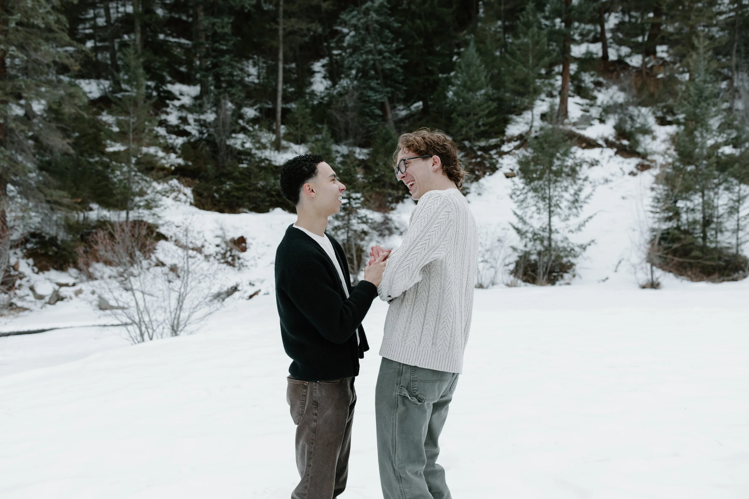 Two people smiling and holding hands in a snowy forest landscape.