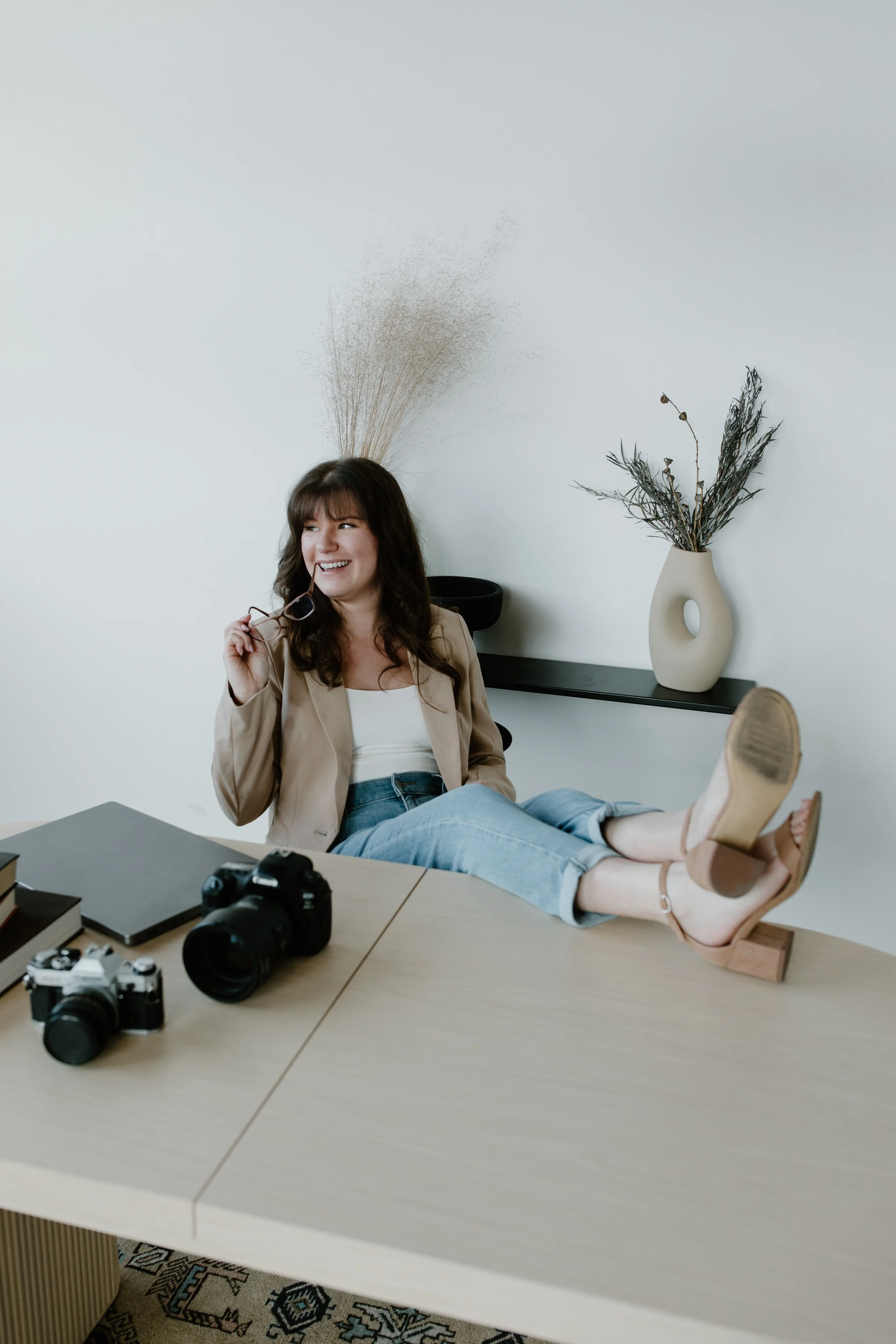 Woman sitting at a desk with her feet on the table, holding glasses, smiling, with cameras on the table.