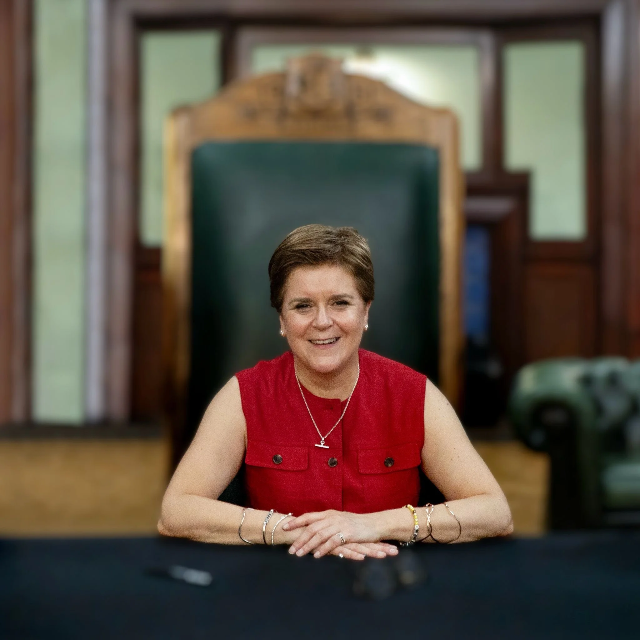 Nicola Sturgeon, the former first minister of Scotland, sits smiling at a table wearing a red top with her arms folded on the desk