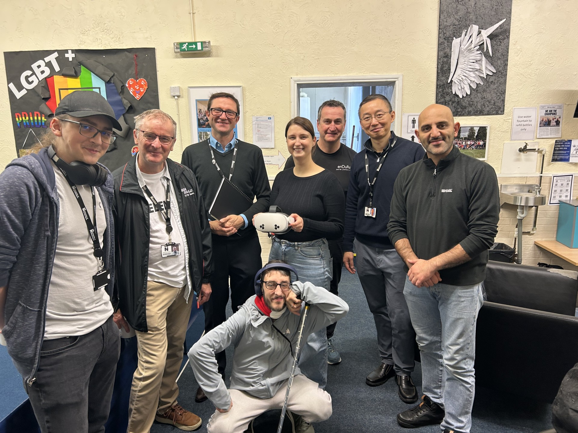A group of people smiling while facing the camera in a room. One is holding a VR headset.