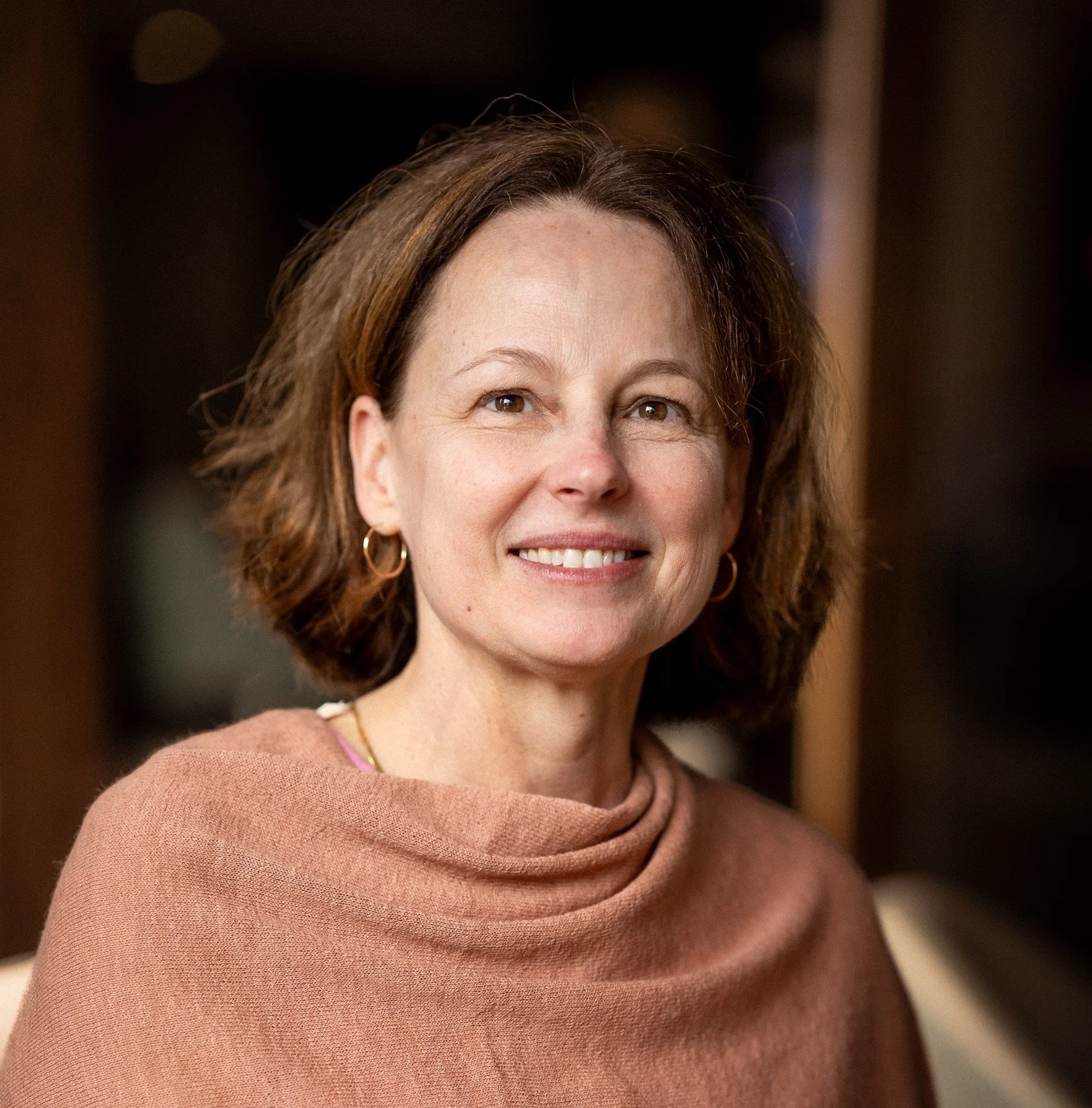 Smiling woman with shoulder-length brown hair, wearing a floral blouse with red, teal, and purple patterns and a purple pendant necklace, standing outdoors with a blurred wooden background.