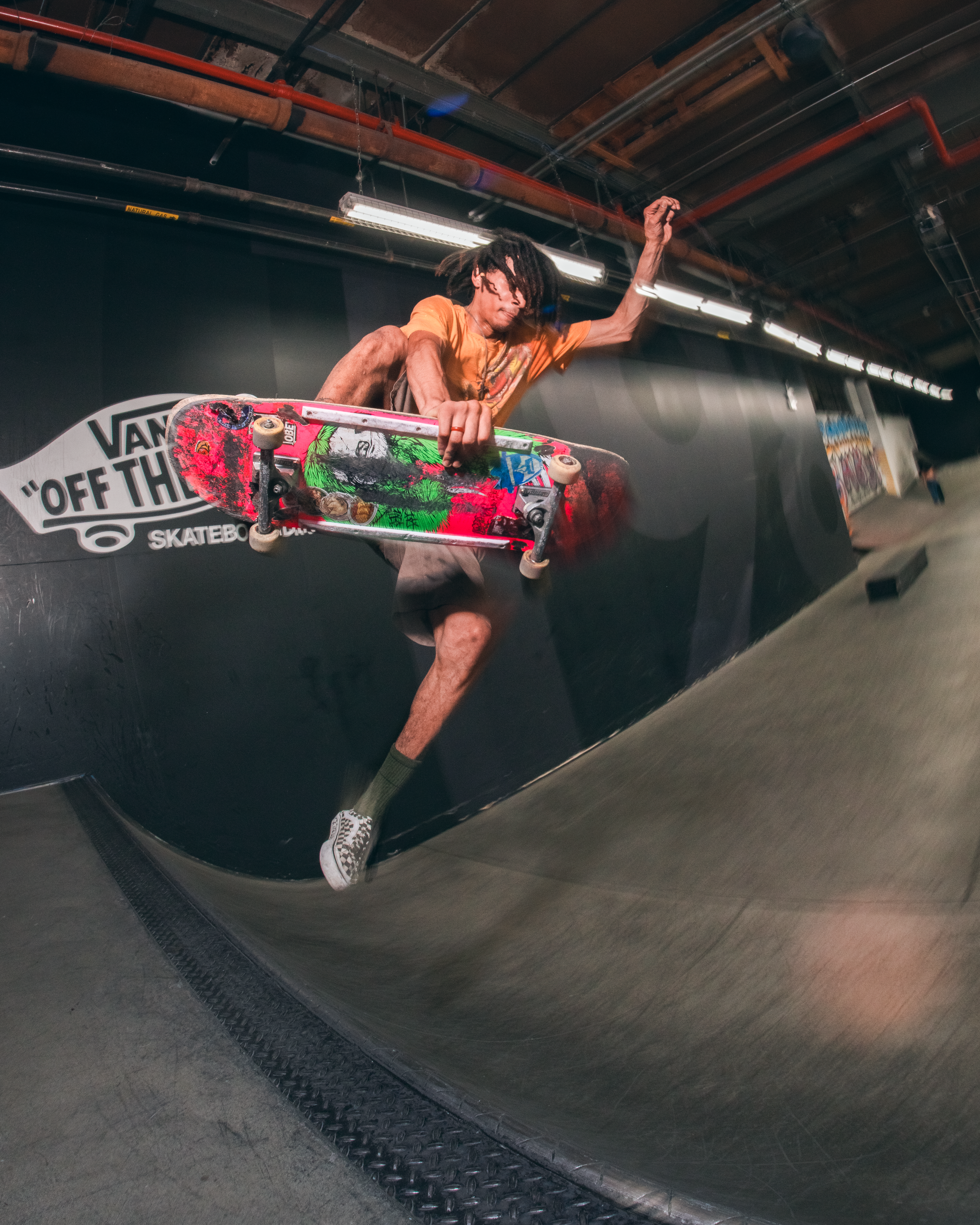 A person with dreadlocks performing a skateboarding trick on a ramp inside an indoor skating facility, reflected in shiny black walls.