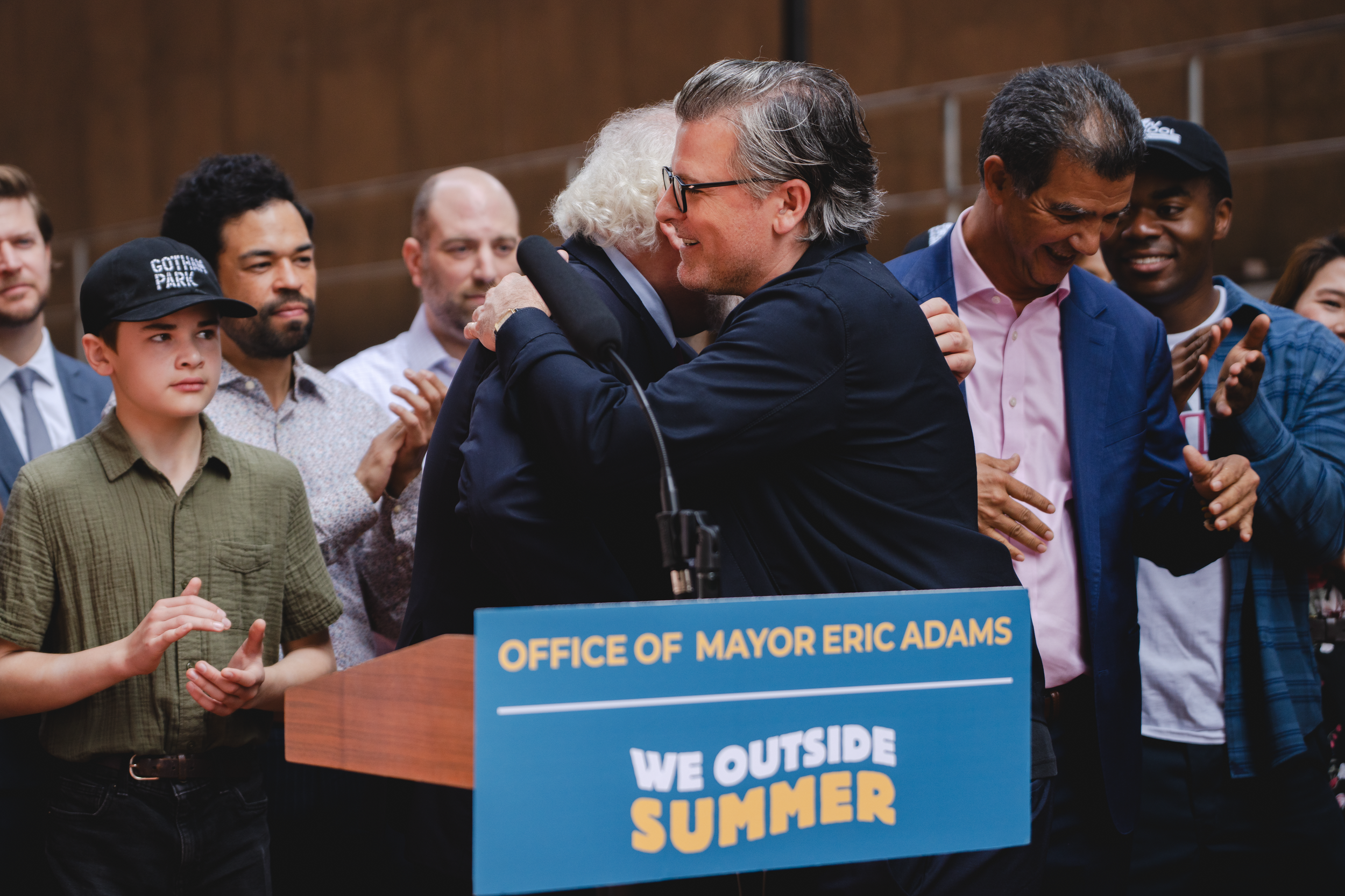 Group of people at a political event, hugging and smiling behind a podium with a sign reading 'Office of Mayor Eric Adams' and 'We Outside Summer' The Banks. Gotham Park Opening