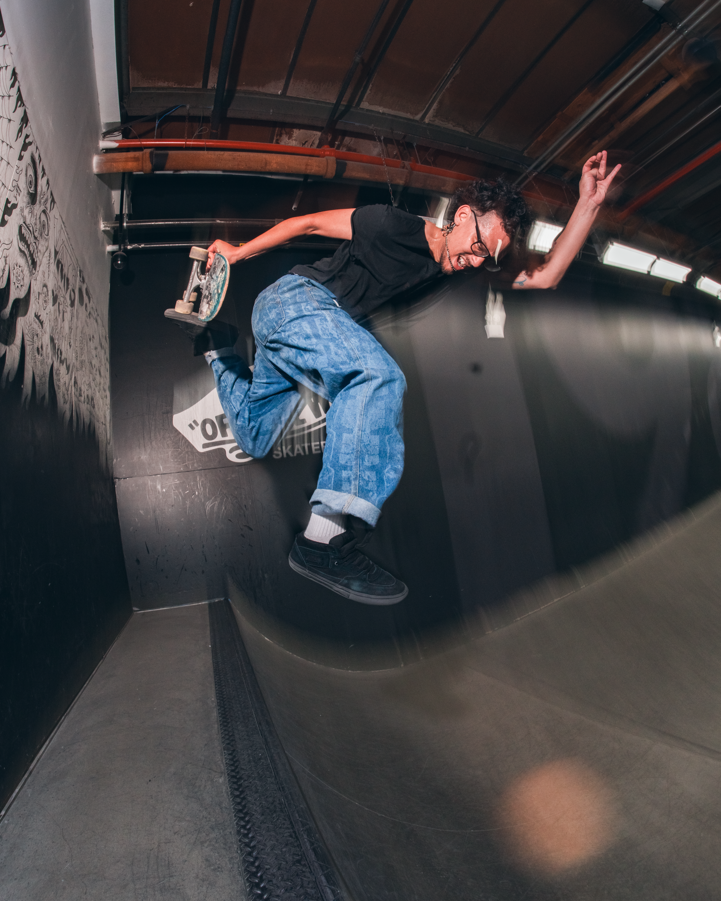 A person skateboarding indoors, caught mid-air with one arm raised and a big smile, wearing glasses, a black t-shirt, and blue jeans.