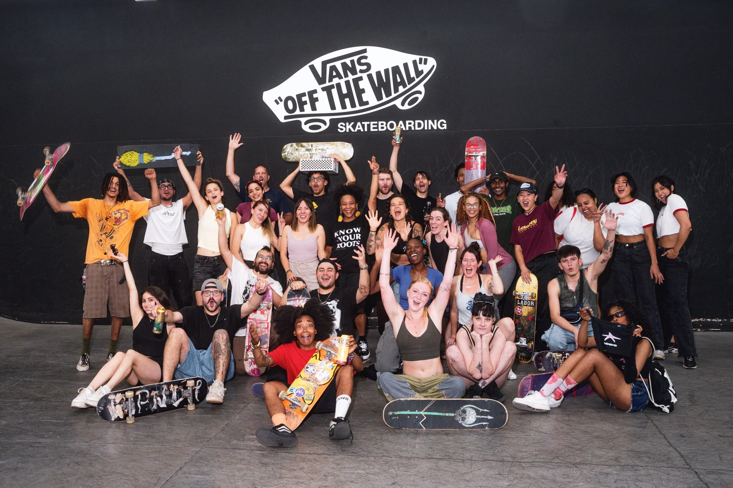 Group of diverse young people smiling and celebrating in front of a Vans skateboarding wall with skateboard decks, some holding drinks and skateboards, posing for a photo.