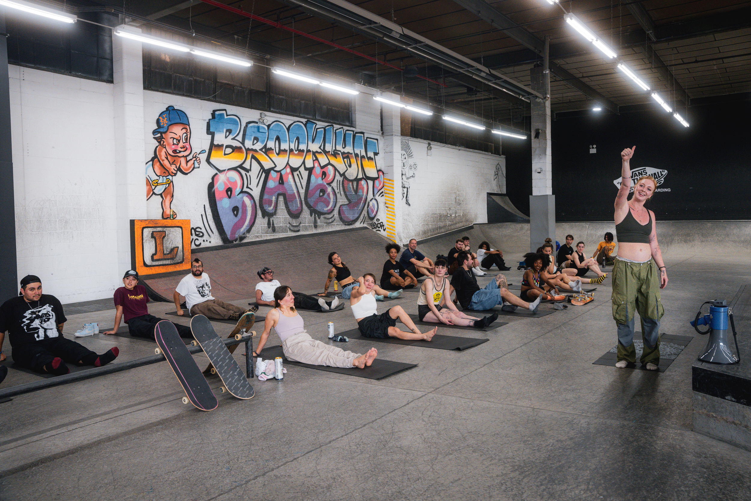 People sitting on the ground with skateboards, participating in a yoga or meditation class in an indoor skatepark with graffiti on the walls.