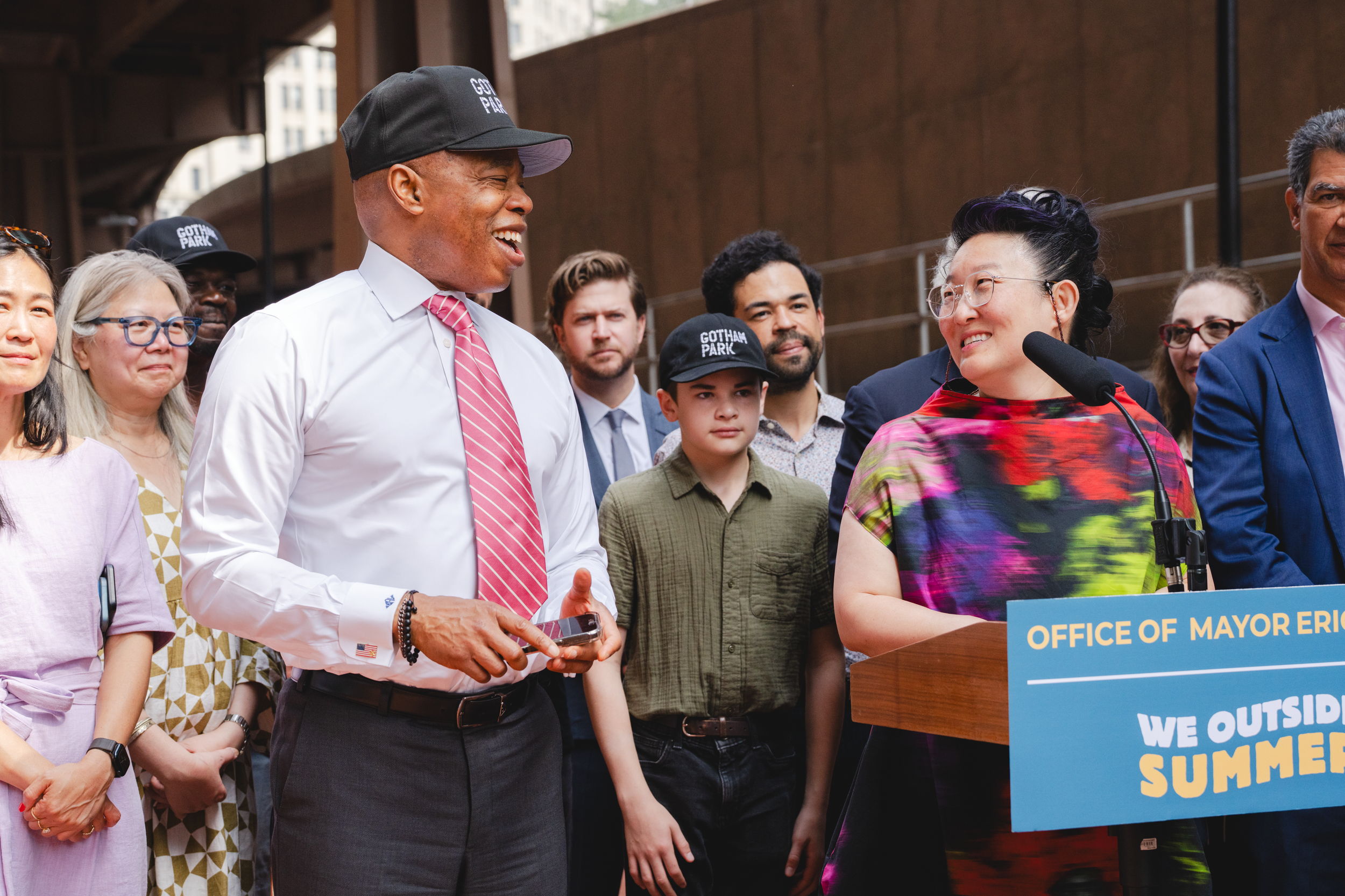 Mayor Eric Adams speaking at a press conference with a group of diverse people, including children and adults, under an overpass, with a sign that reads 'Office of Mayor Eric Adams' and slogans about summer programs. The Banks. Gotham Park Opening.