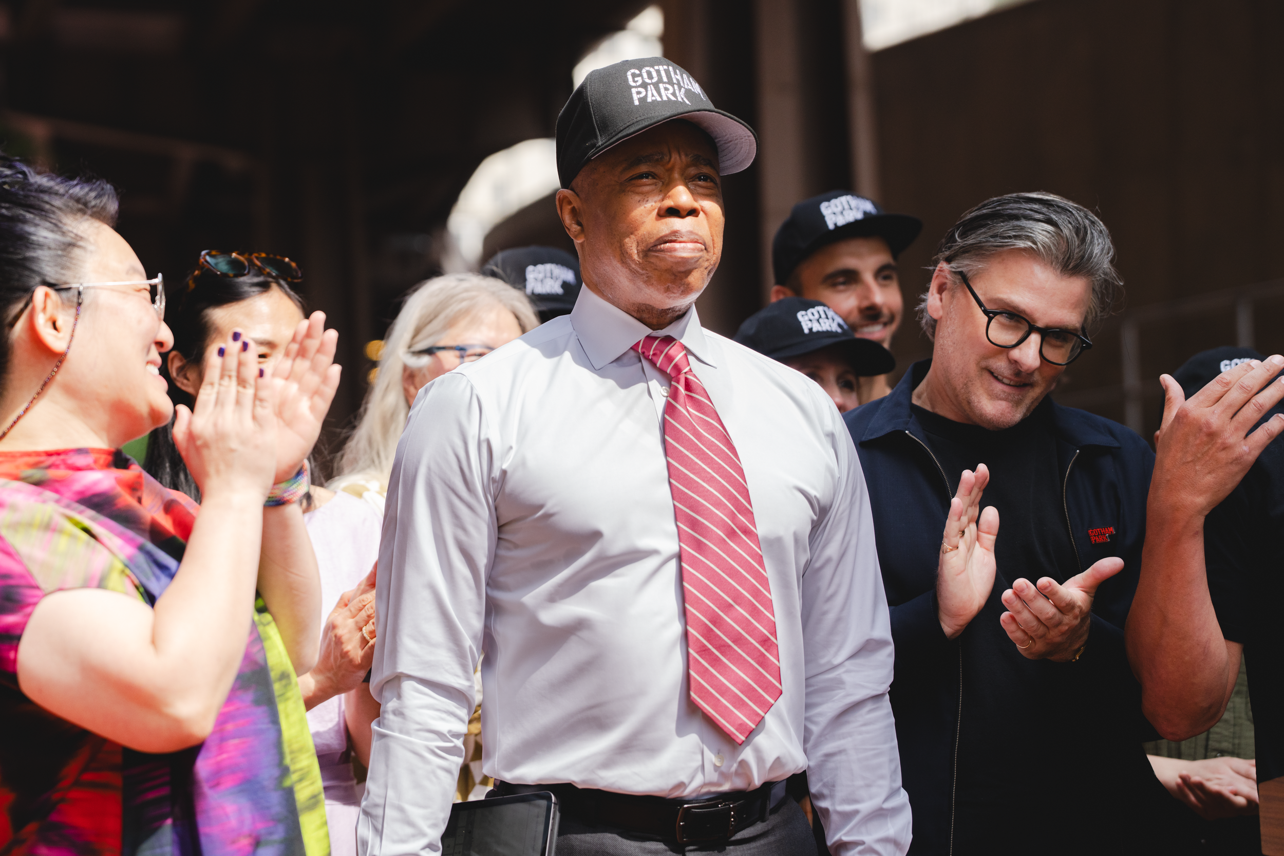 A group of people, including a man in a white shirt, red striped tie, and a Gotham Park cap, standing among others who are clapping and smiling, at an outdoor event. The Banks. Gotham Park Opening. Eric Adams