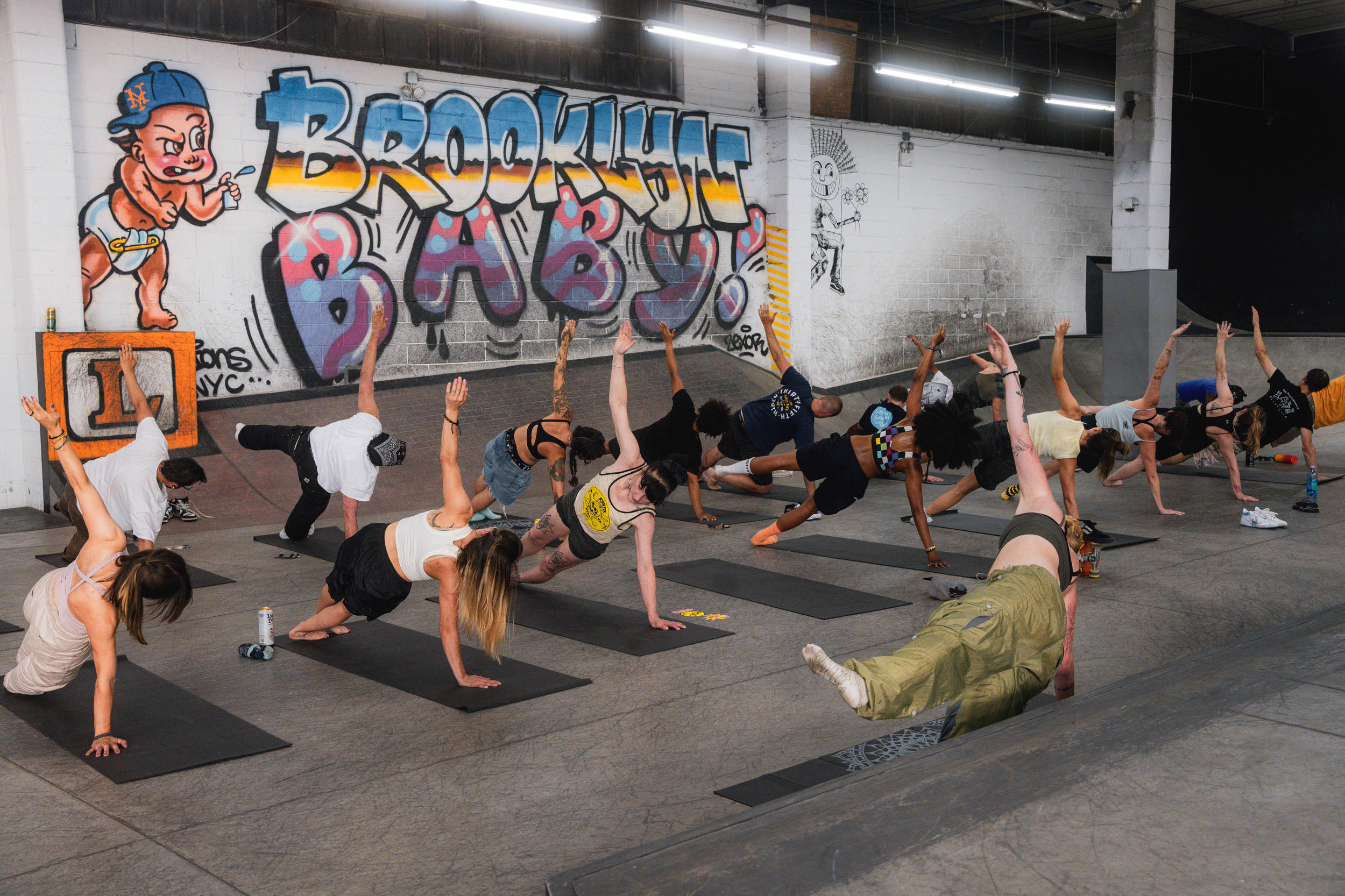A group of people participating in a yoga class in an industrial-style gym with graffiti artwork on the wall. The participants are in a side plank pose, with some looking at the instructor or the wall, on black mats. The graffiti includes a cartoon b