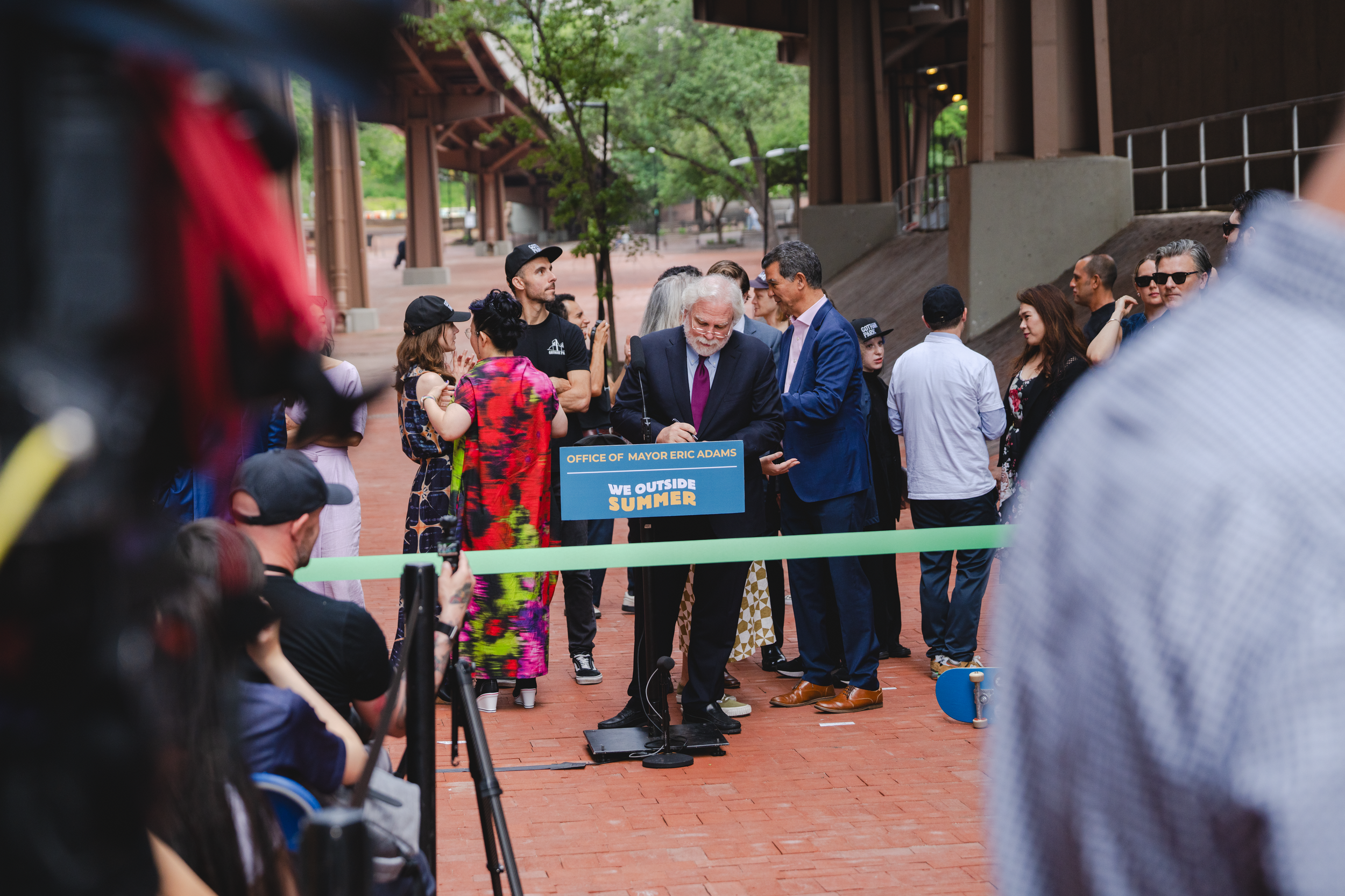 A group of people gathered in an outdoor urban setting for a ribbon-cutting event, with a man in a suit signing a document at a podium marked 'Office of Mayor Eric Adams' and a sign that reads 'We Outside Summer'. The Banks. Gotham Park Opening