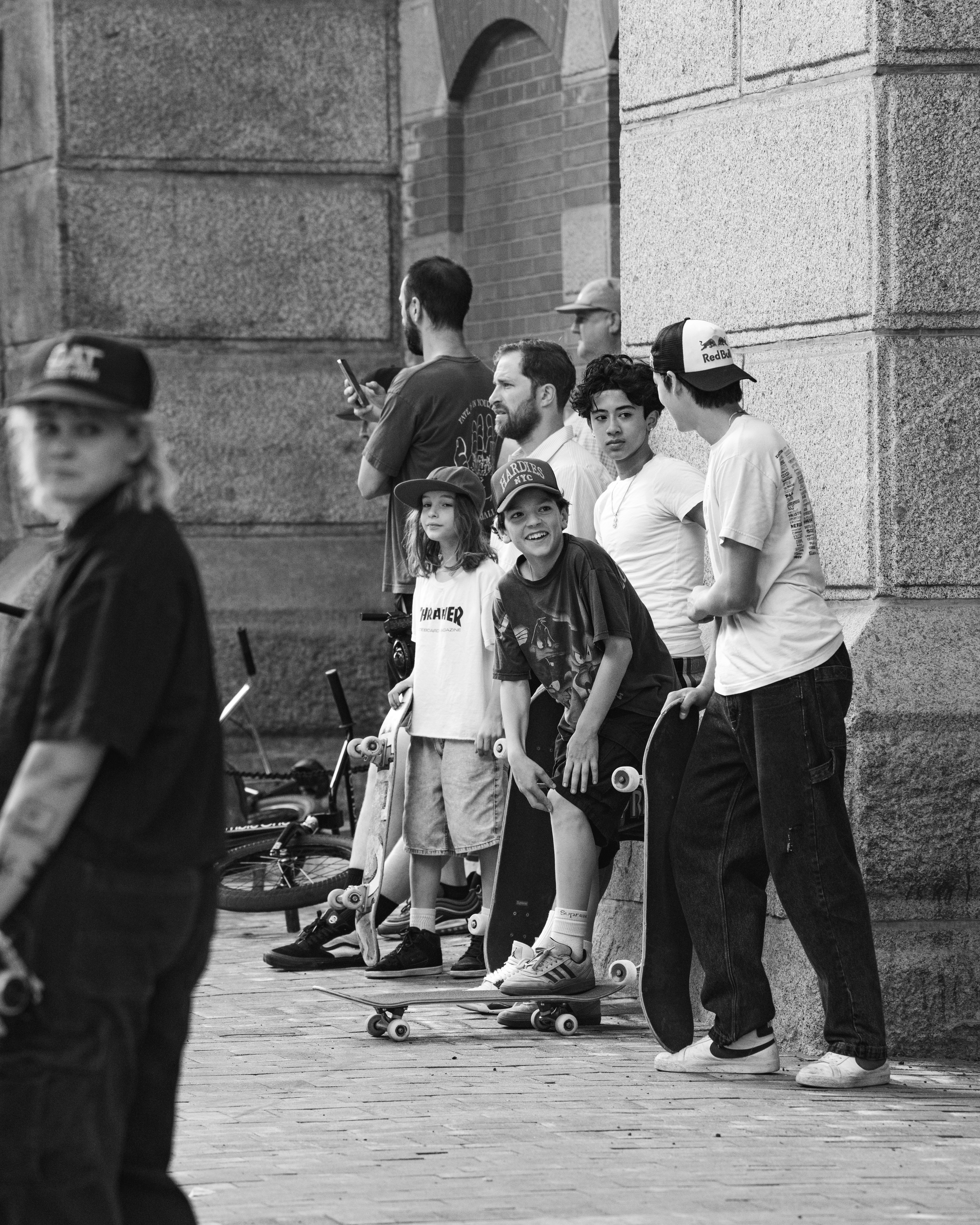 A group of young people with skateboards standing and talking on a city sidewalk against a stone wall, some looking at their phones, in black and white.